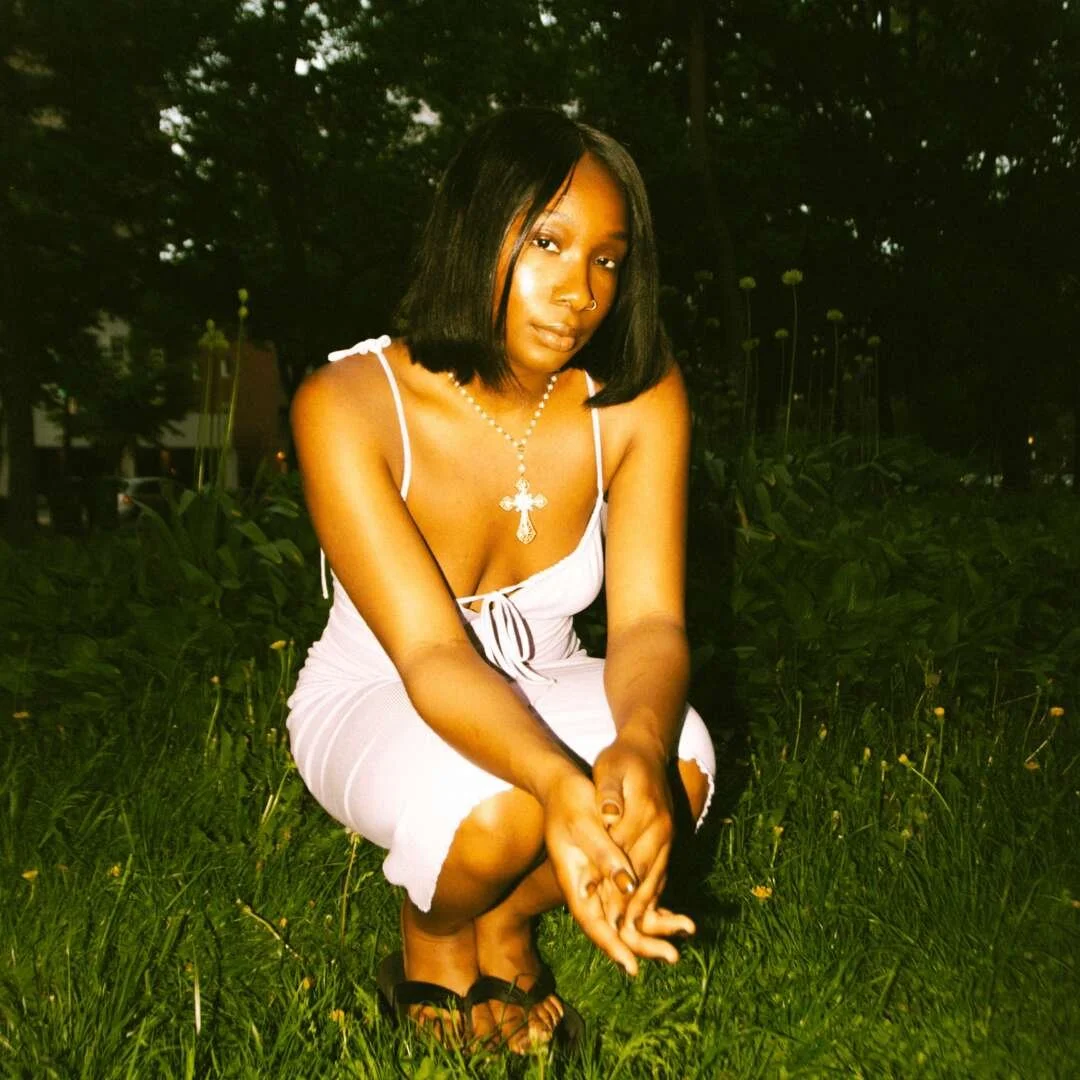 A woman with shoulder-length black hair, wearing a white dress with spaghetti straps and sandals, is squatting on grass in a park at dusk, with trees in the background and a silver cross necklace.