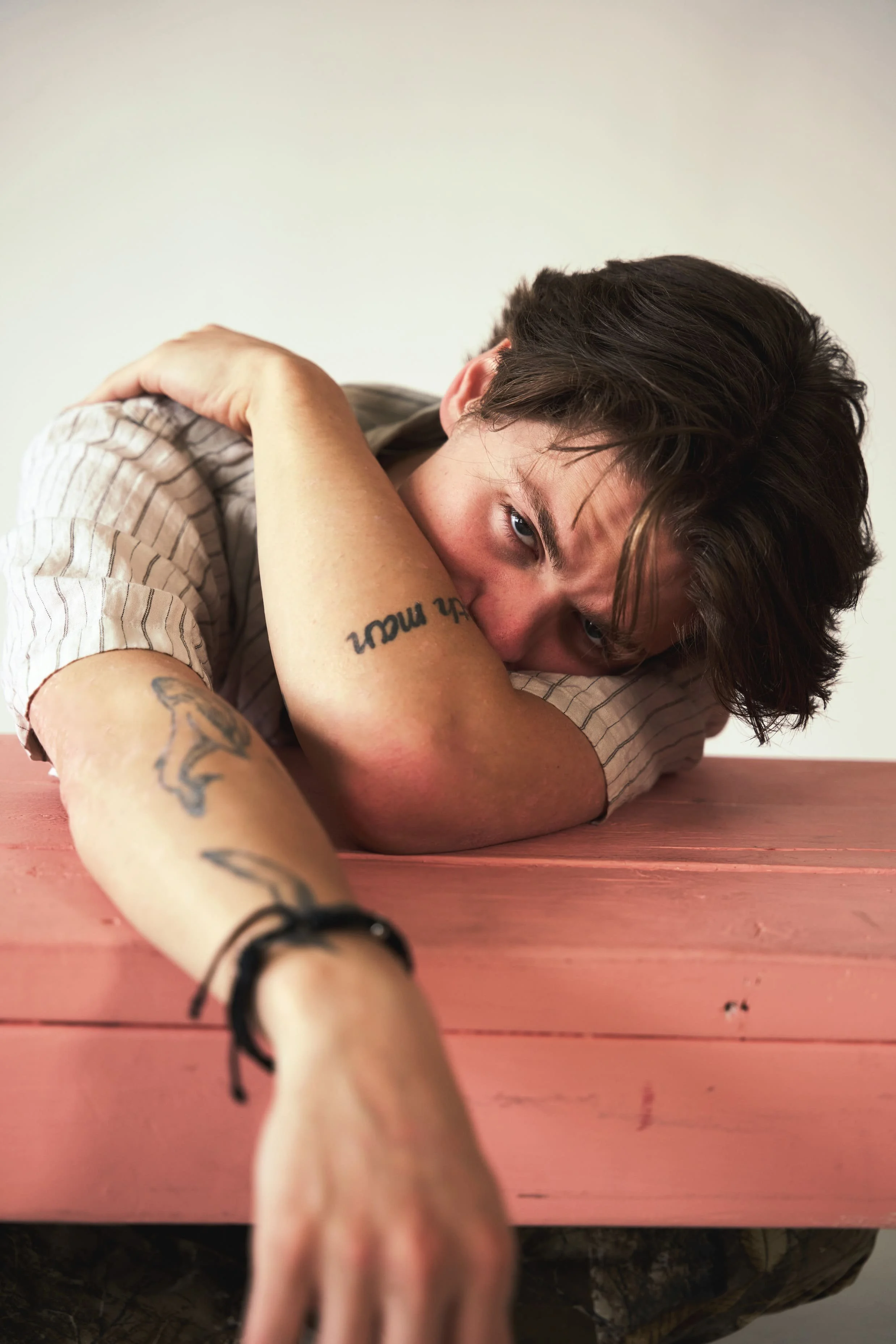A young man with dark, wavy hair sitting on a wooden chair against a plain, light gray background. He is dressed in a dark sweatshirt with a graphic design, oversized dark green cargo pants, and sneakers. His posture is relaxed, with one leg crossed over the other, and he appears to be gazing forward.