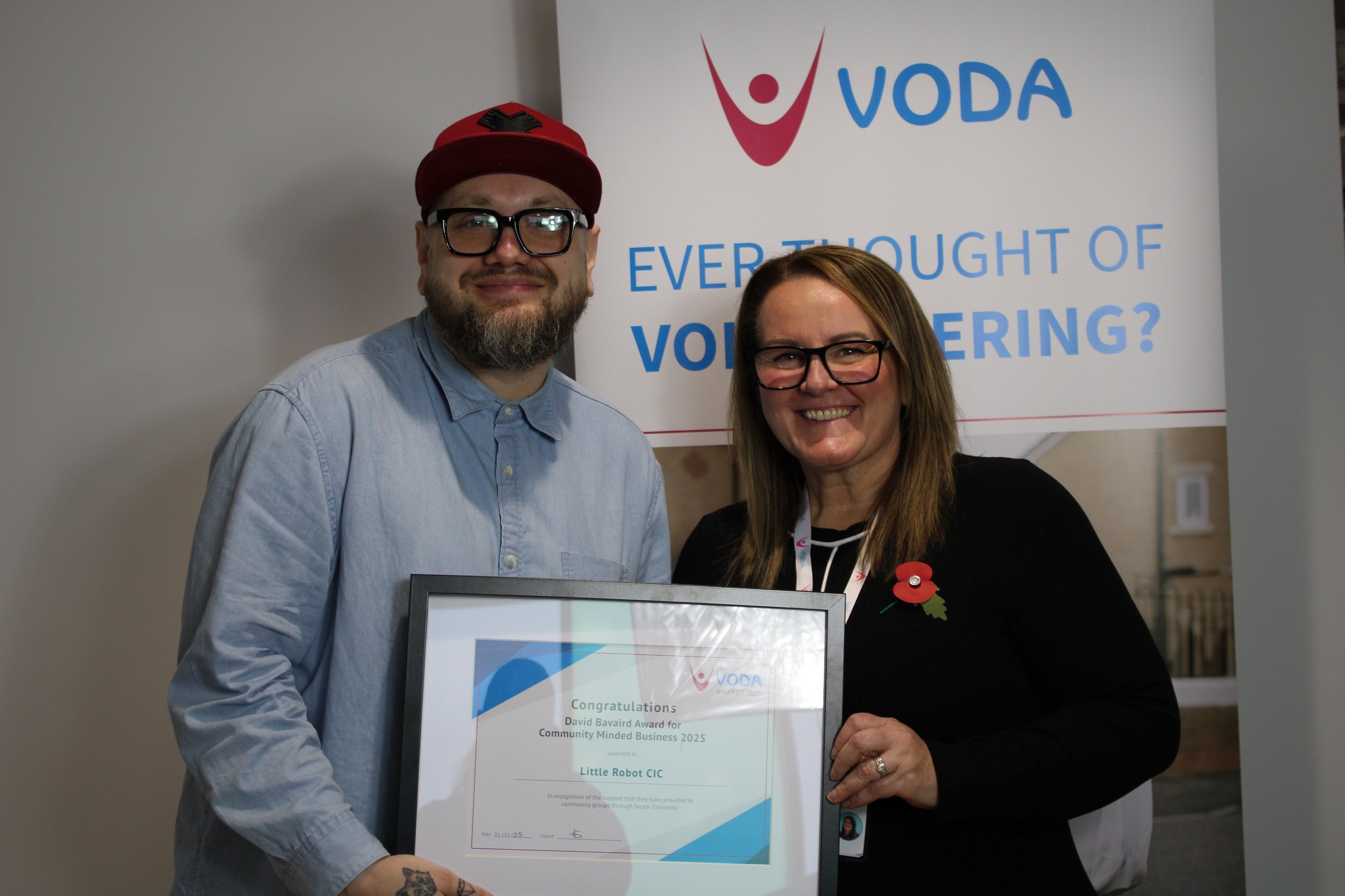 A man and woman smiling and holding a framed award certificate at a VODA event with a banner in the background that reads 'EVER THOUGHT OF VOICE SCHEDULING?'