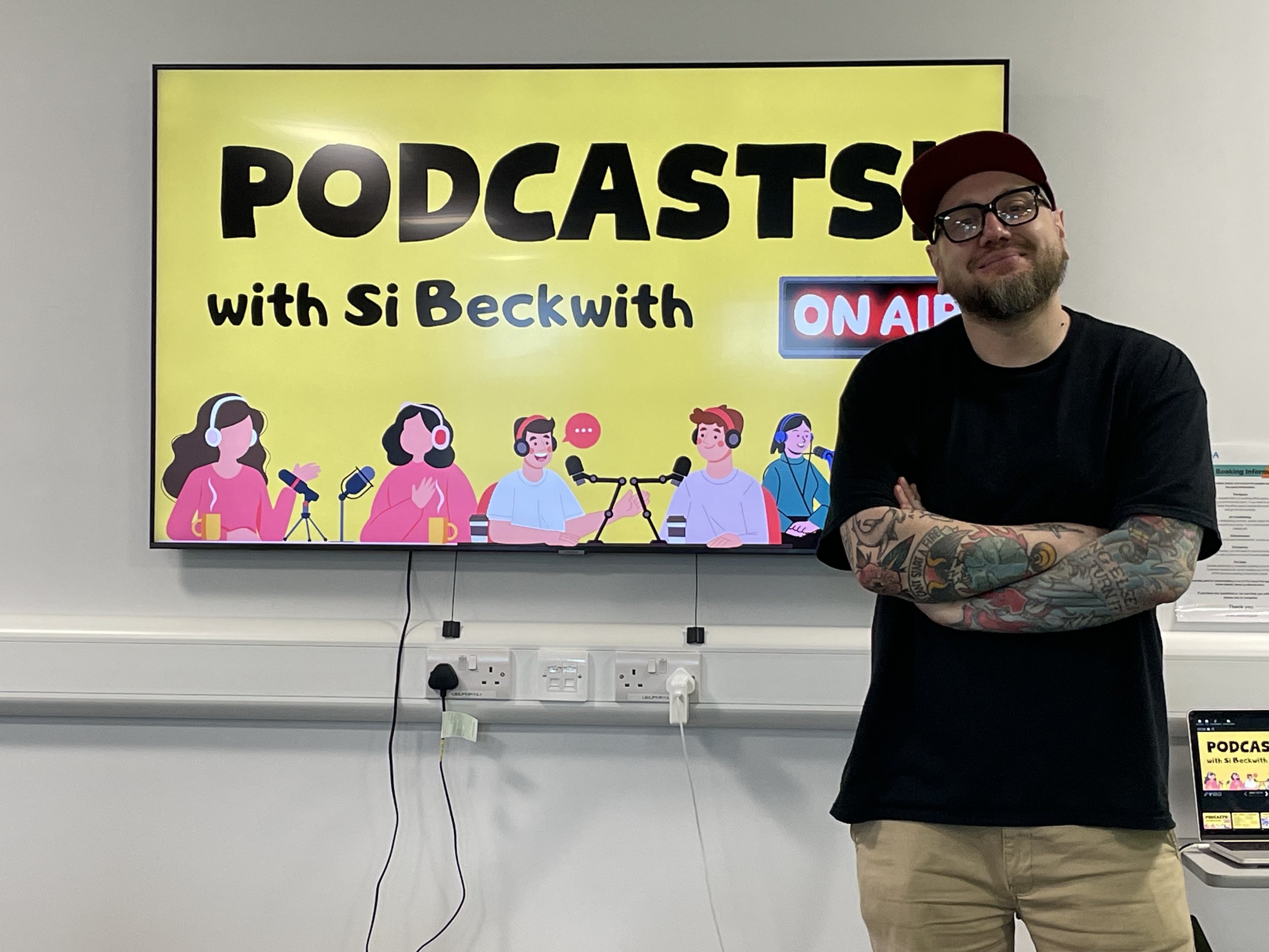 Man with arm tattoos standing in front of a screen advertising a podcast with cartoon illustrations of people, a microphone, and an 'On Air' sign.