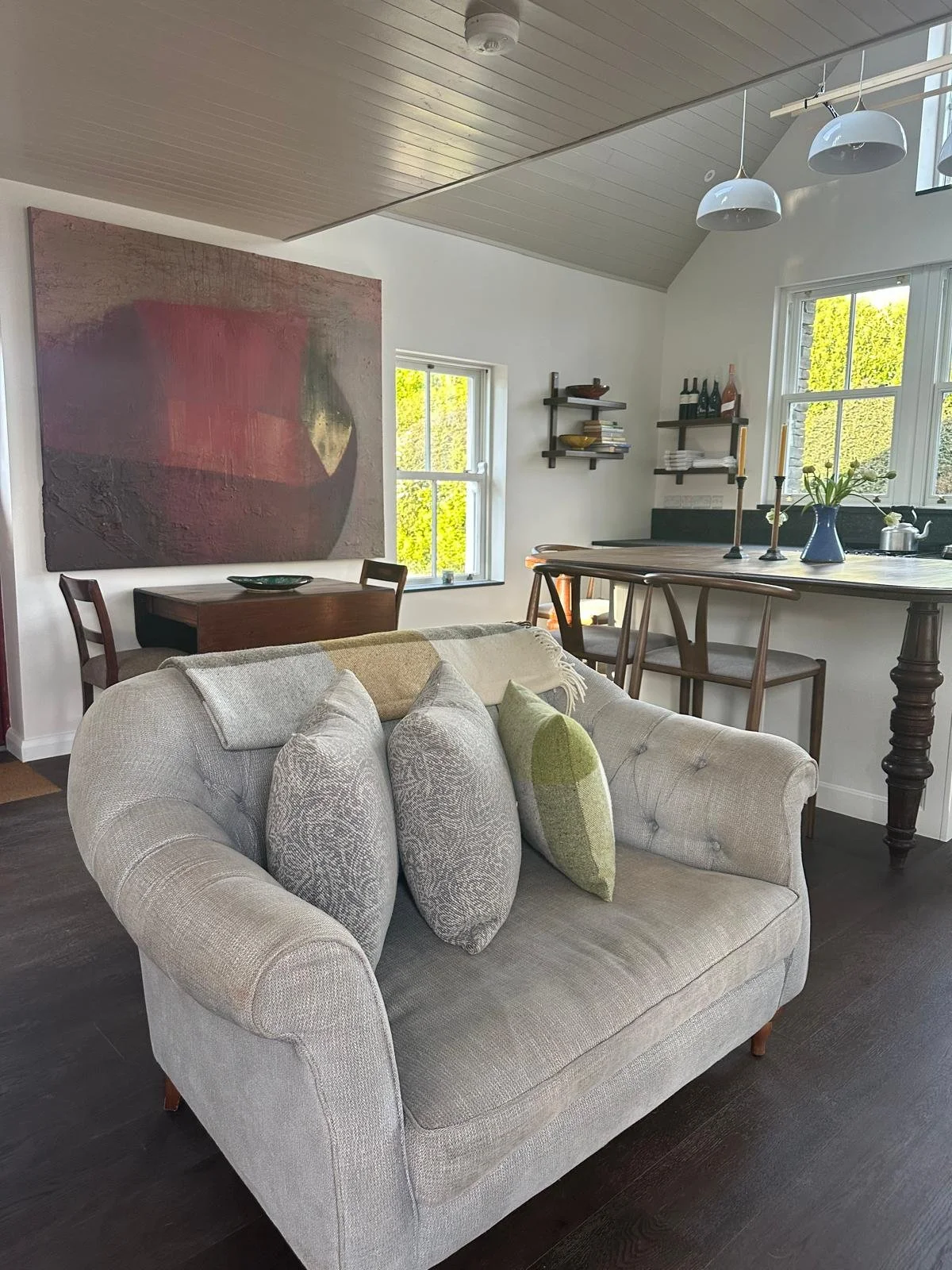 A cozy living room with a beige tufted sofa adorned with patterned and solid-colored pillows. In the background, a dark wooden dining table with two chairs, a sideboard under the window, and shelves with books and decorative items. Large windows allo