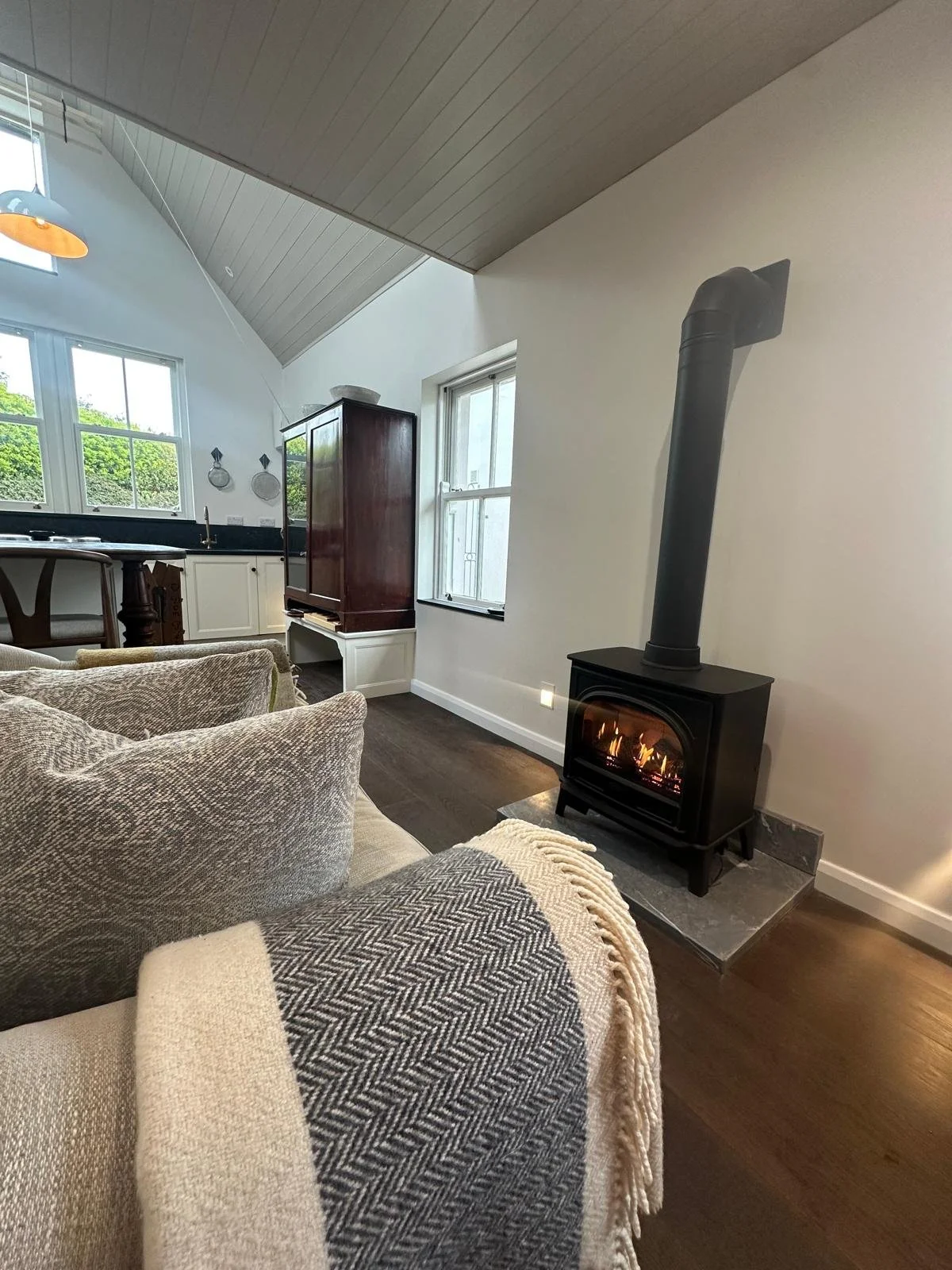 Living room with a wood-burning stove, couches with throw blankets, large windows, and a kitchen area in the background.