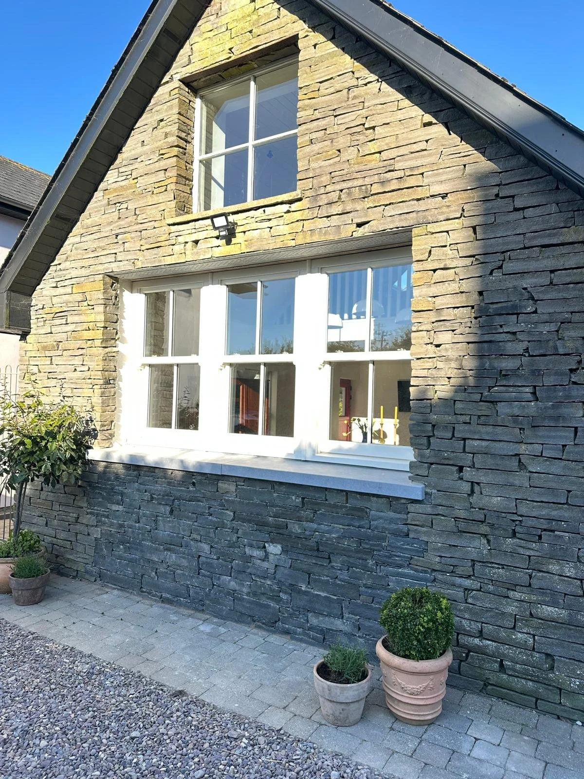 A house exterior with a stone facade, large rectangular windows with white frames, and a small garden with potted plants in front, under a clear blue sky.
