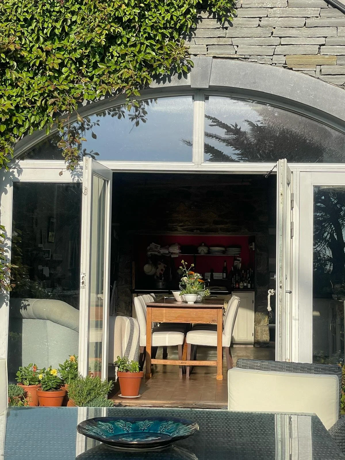 Open glass doors of a house revealing a cozy kitchen and dining area with a wooden table, white chairs, and potted plants outside.