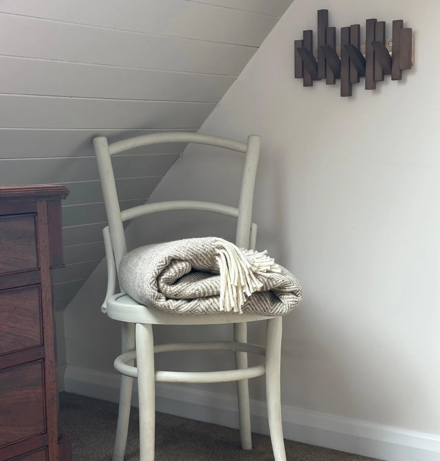 A white wooden chair with a folded beige blanket on it, placed against a wall with angled ceiling, next to a wooden dresser and a dark modern wall art piece.