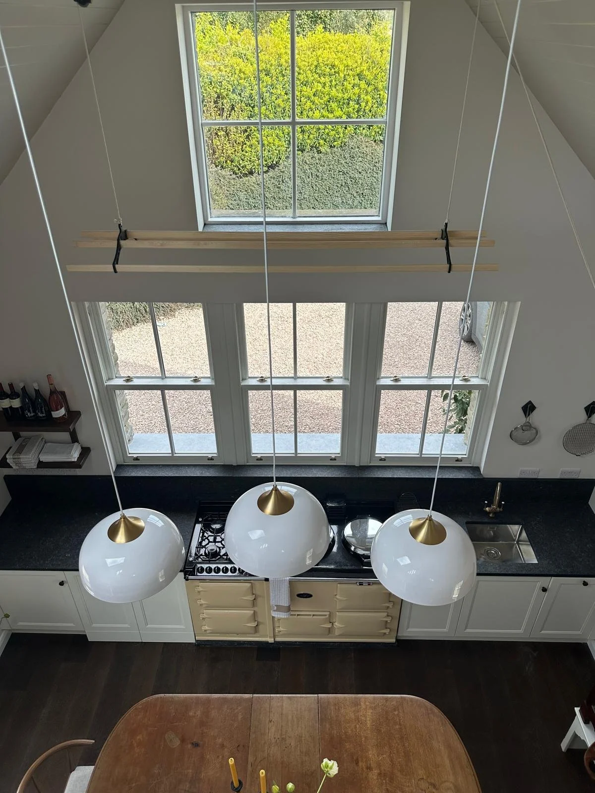 Overhead view of a modern kitchen with a black countertop, white cabinets, and three large hanging pendant lights with white shades and gold accents. Behind the counter are large white-framed windows and a door with glass panes, letting in natural li