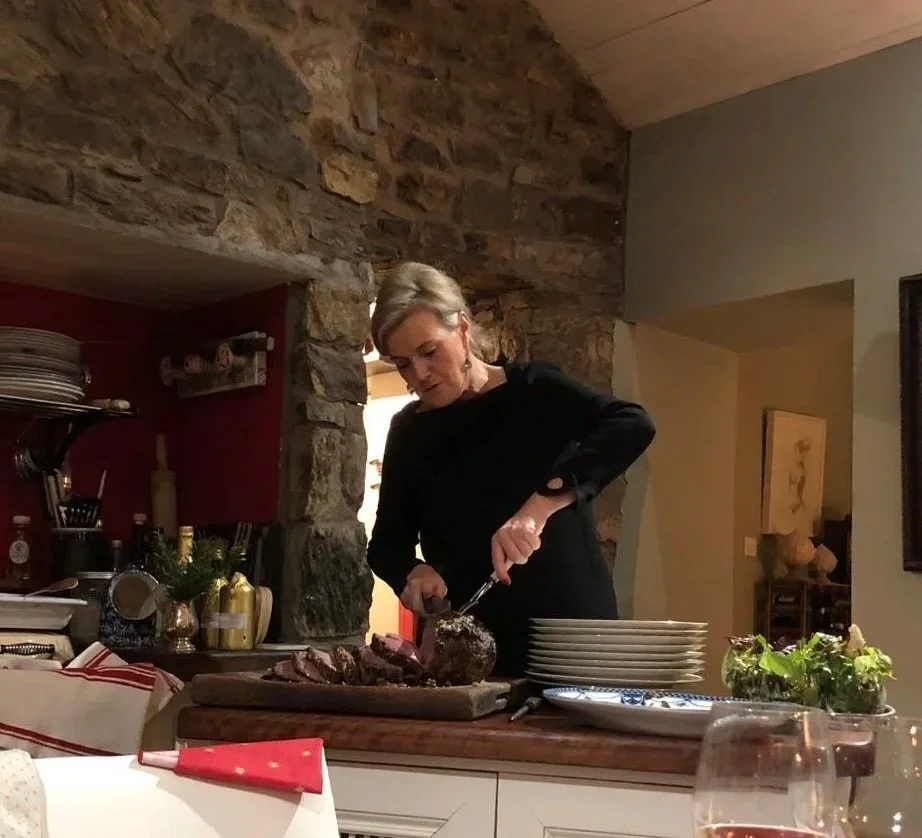 A woman with gray hair cooking or preparing food in a kitchen, standing behind a wooden counter with plates, a mixing bowl, and a pie on a cutting board.