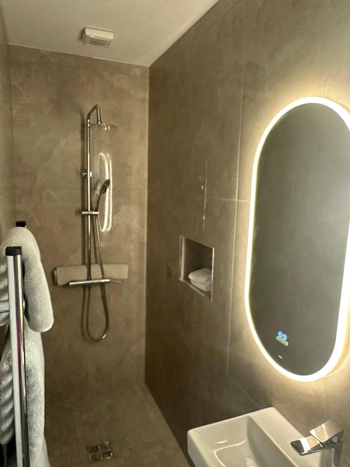 Modern bathroom with a showerhead, a backlit mirror, a towel rack, a soap dish, and beige tiled walls and floor.