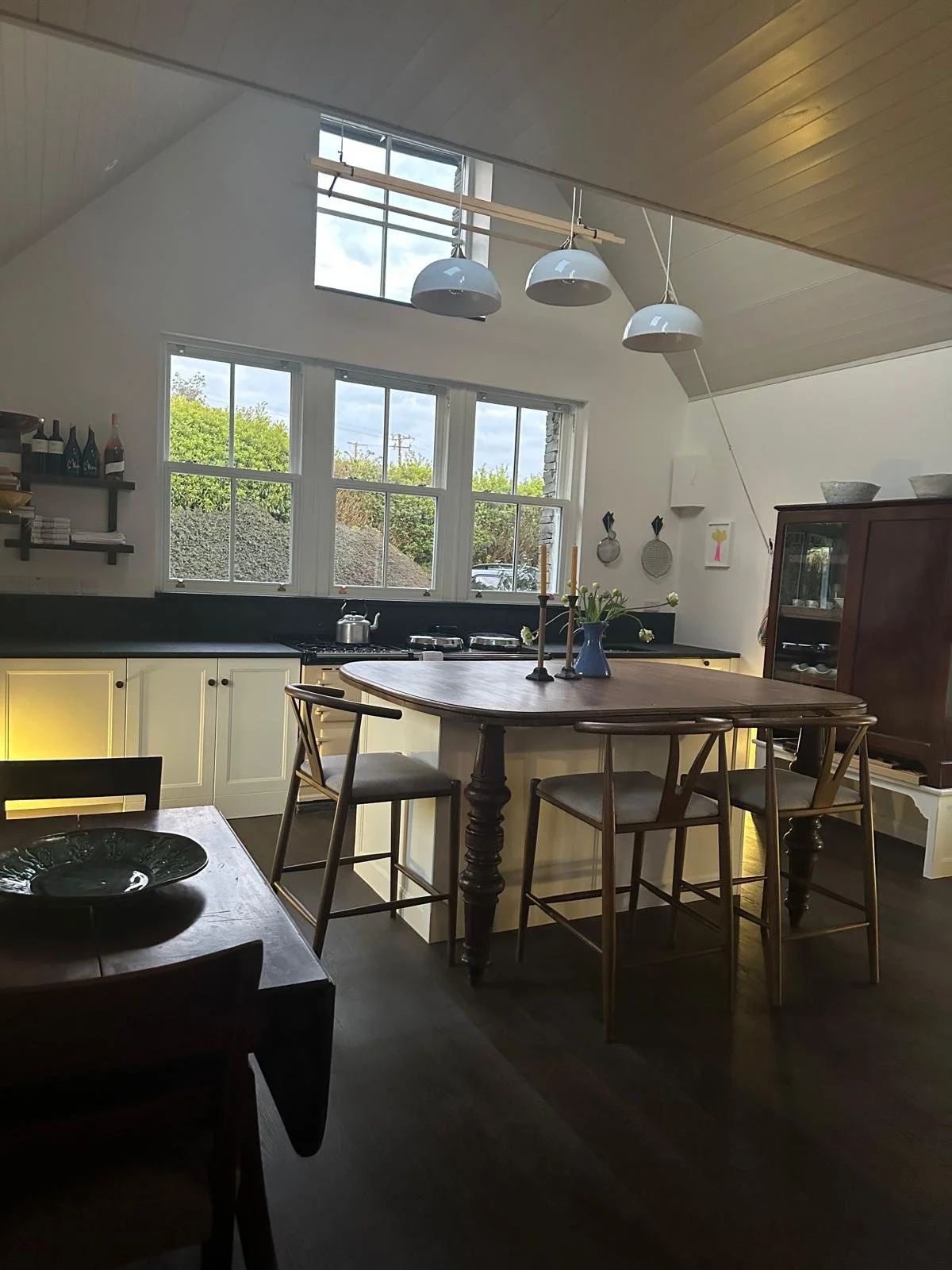 A cozy kitchen featuring white cabinetry, a black countertop, and a central wooden dining table with chairs. Large windows let in natural light, and decorative items such as bottles, candles, and a vase with flowers adorn the space.