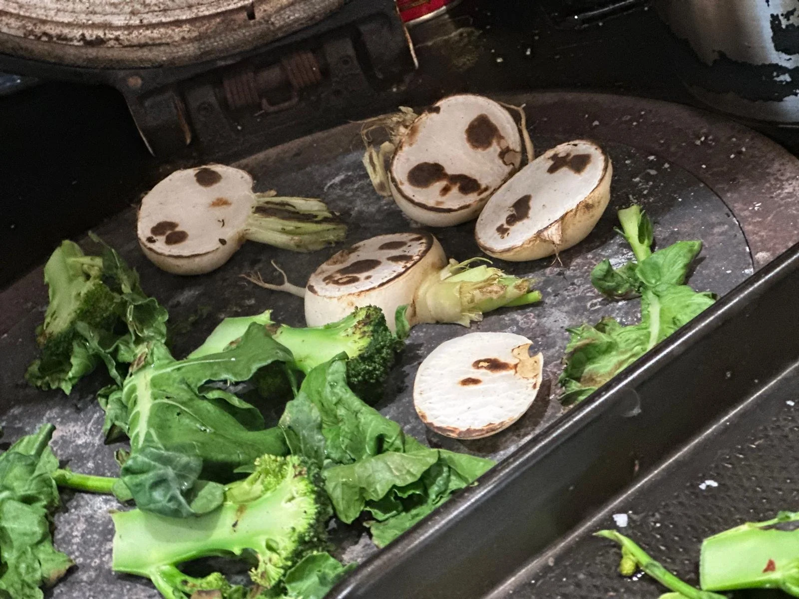 Vegetables on a griddle, including sliced turnips and broccoli florets, for cooking.