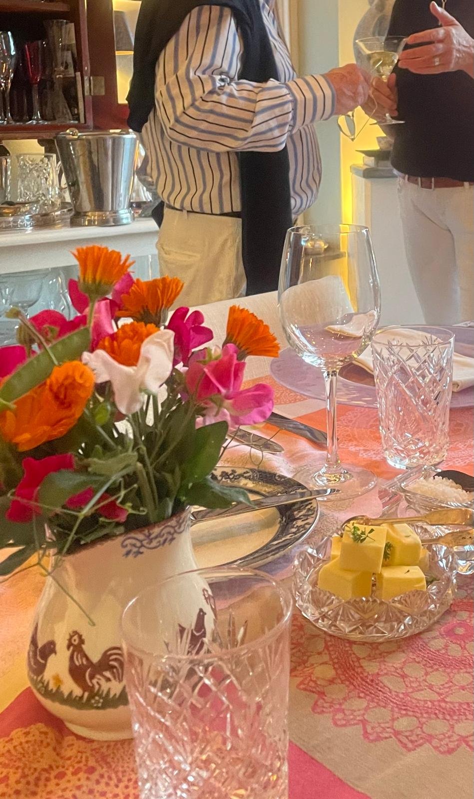A table with a flower arrangement, wine glass, water glass, plate of butter, and cutlery, with two people standing in the background holding drinks.