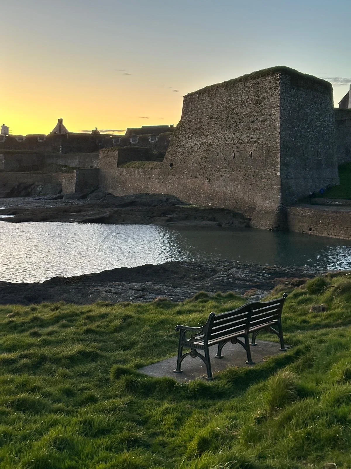 Historic stone fort or castle overlooking water at sunset, with a grassy area and a black metal bench in the foreground.