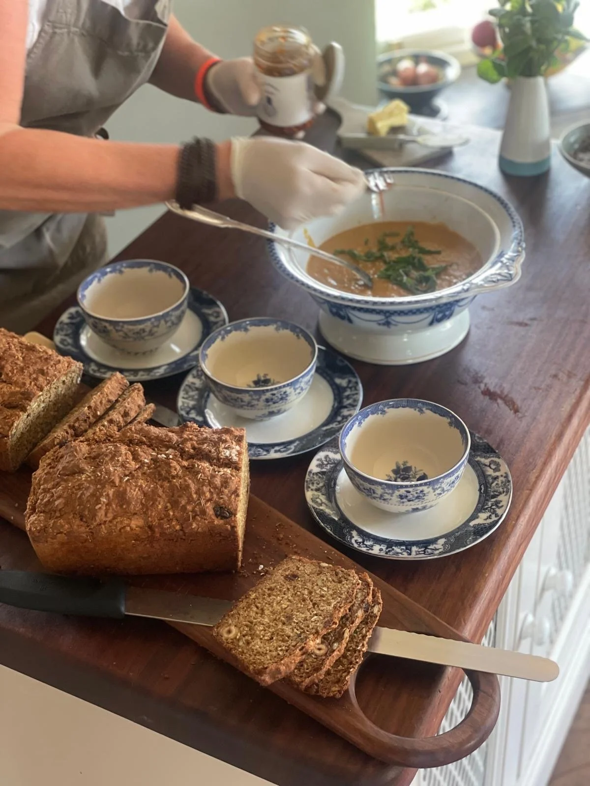 Person preparing a bowl of soup on a wooden table, with sliced bread, tea cups, and a jar of spread or honey. There are flowers in a white vase in the background.
