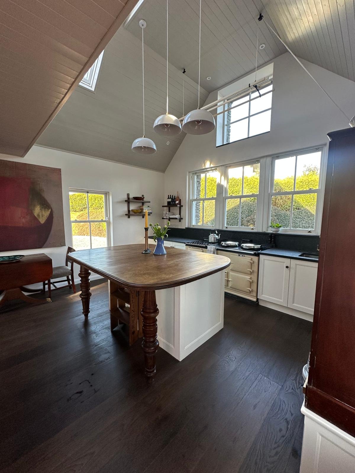 A kitchen with a high peaked ceiling, large windows, white cabinets, black countertop, and a wooden island table with a vase of tulips and a candlestick. There are hanging pendant lights and wall-mounted shelves with bottles and dishes.