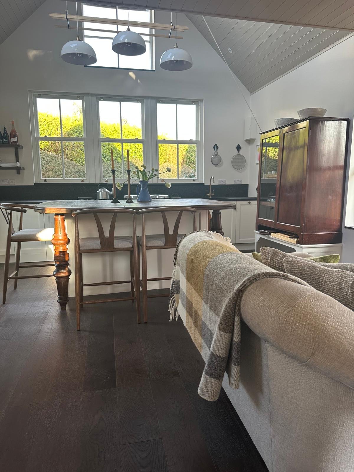 A cozy kitchen and living room area with a large window overlooking greenery, a wooden dining table with chairs, a sofa with a plaid throw, and decorative items like candles and a vase of flowers.
