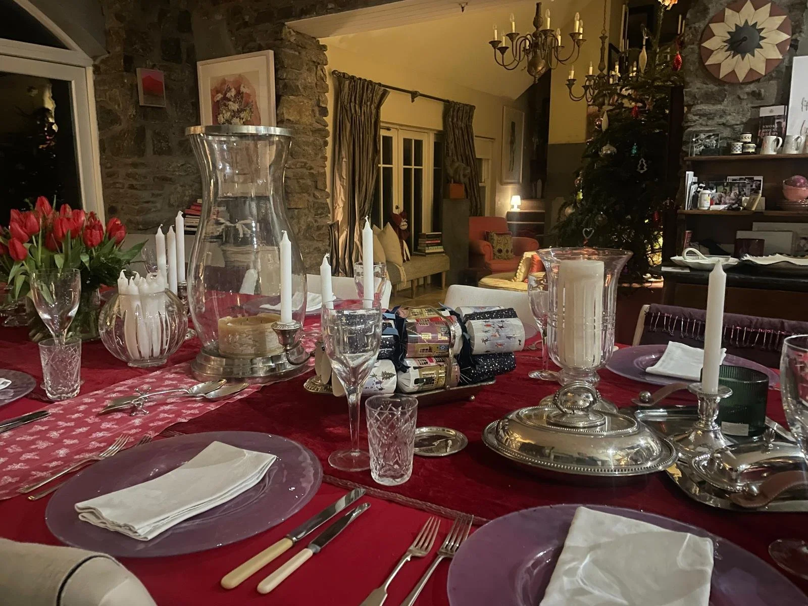 A decorated dining table set for a holiday meal, with a red tablecloth, white napkins, candles, wine glasses, and presents, in a cozy home with a Christmas tree in the background.