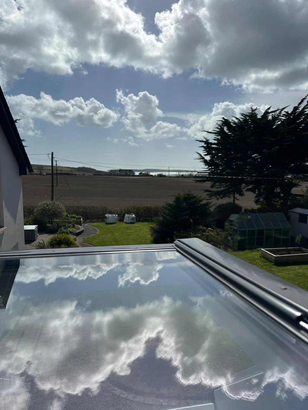 Cloudy sky reflected on glass roof of a car, with a view of a backyard, trees, a greenhouse, and a farmland in the distance.