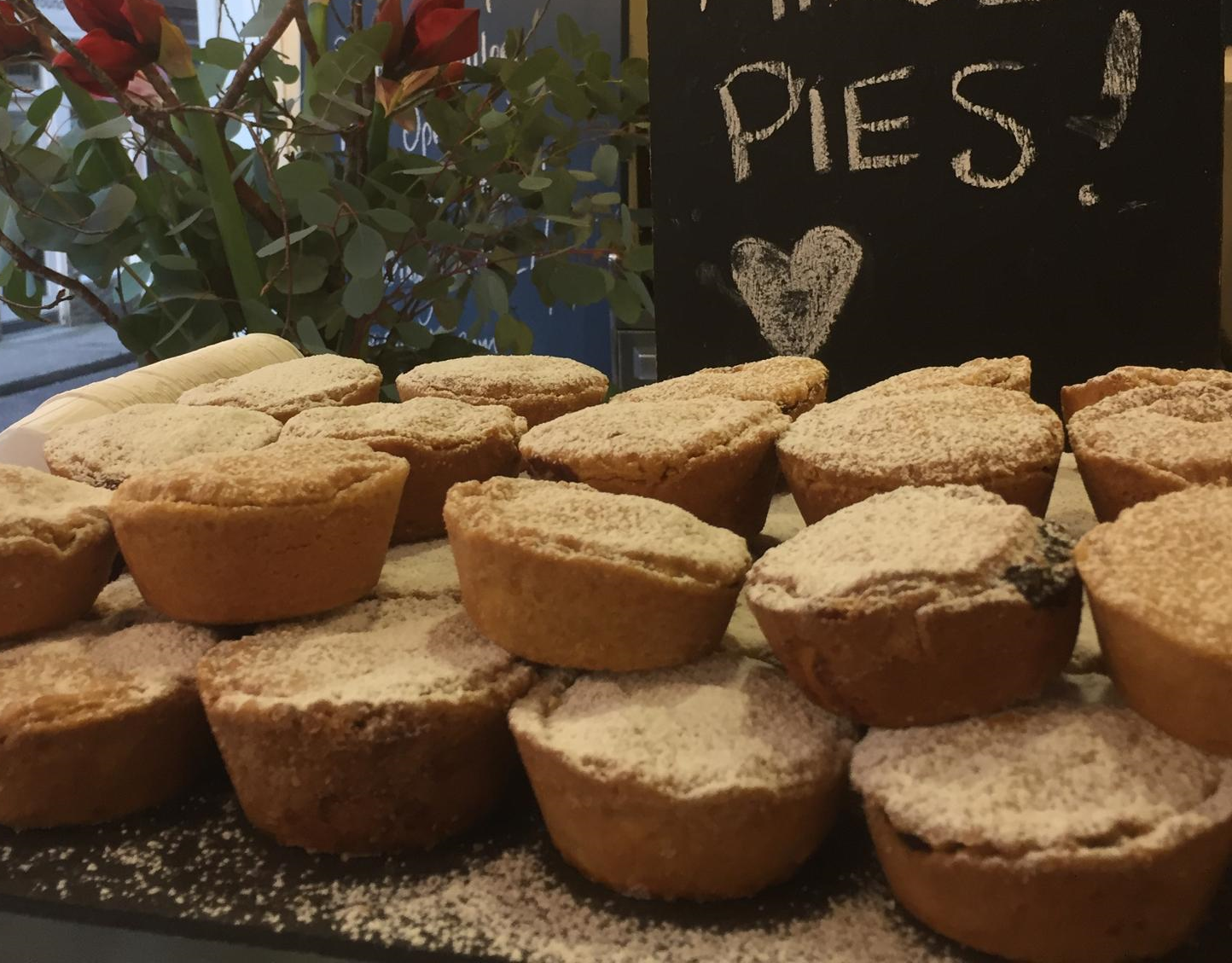Multiple powdered sugar-topped mince pies arranged on a tray with a small chalkboard sign behind them that says 'Pies?' with a heart drawing.