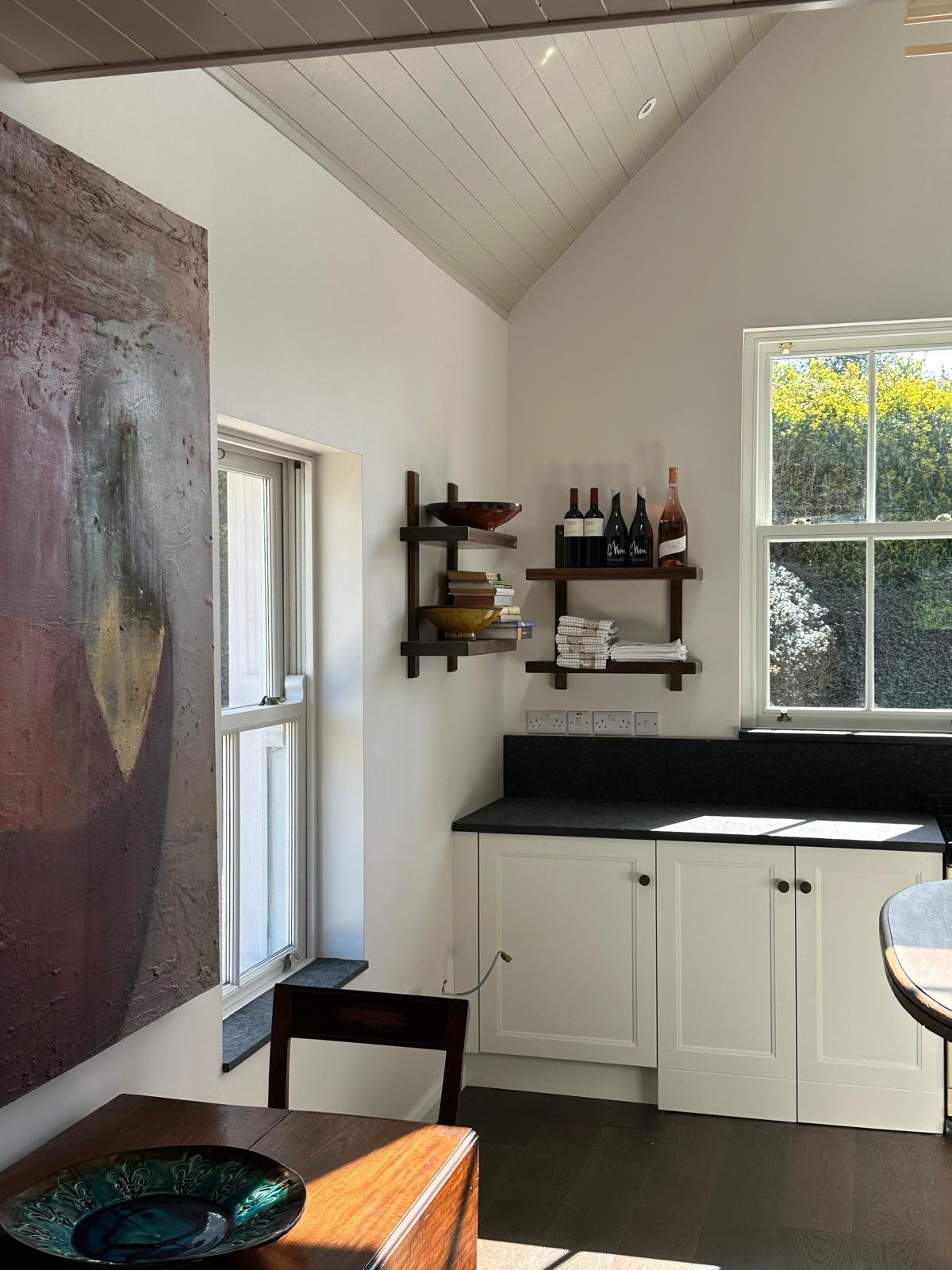 A kitchen corner with white cabinetry, black countertop, wooden shelves holding wine bottles and folded towels, a window with trees outside, and a wall-mounted artwork.