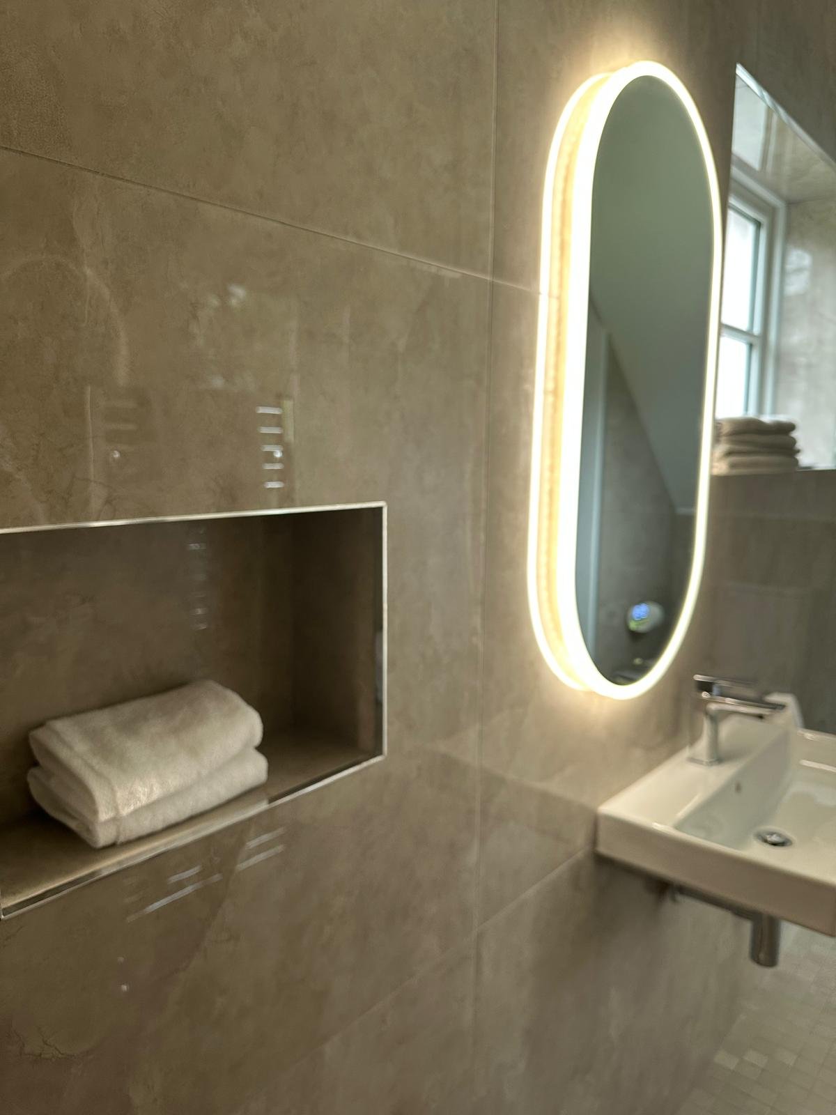 Modern bathroom with beige tiled walls, a backlit oval mirror, a small rectangular sink with a chrome faucet, and a window with folded towels in the background.