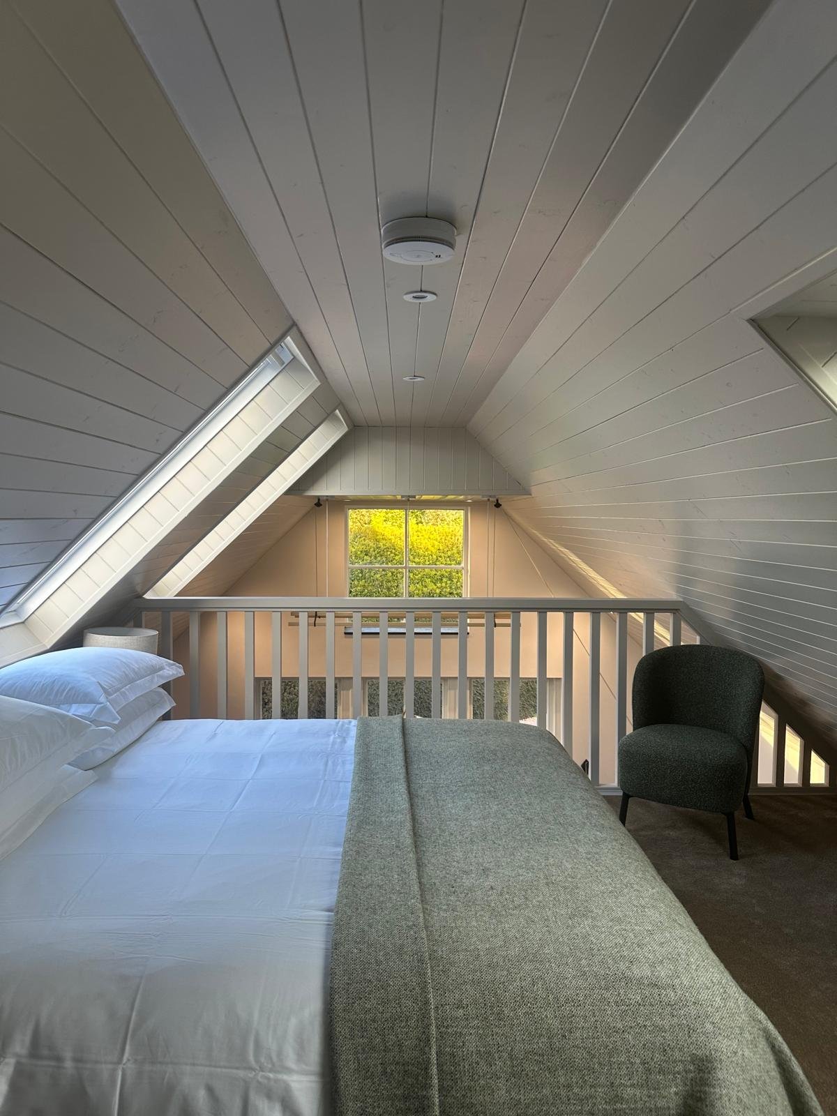 A cozy bedroom with sloped white wooden ceiling, a large bed with white sheets and pillows, a gray blanket, a black armchair, a white lampshade, and a window showing green foliage.