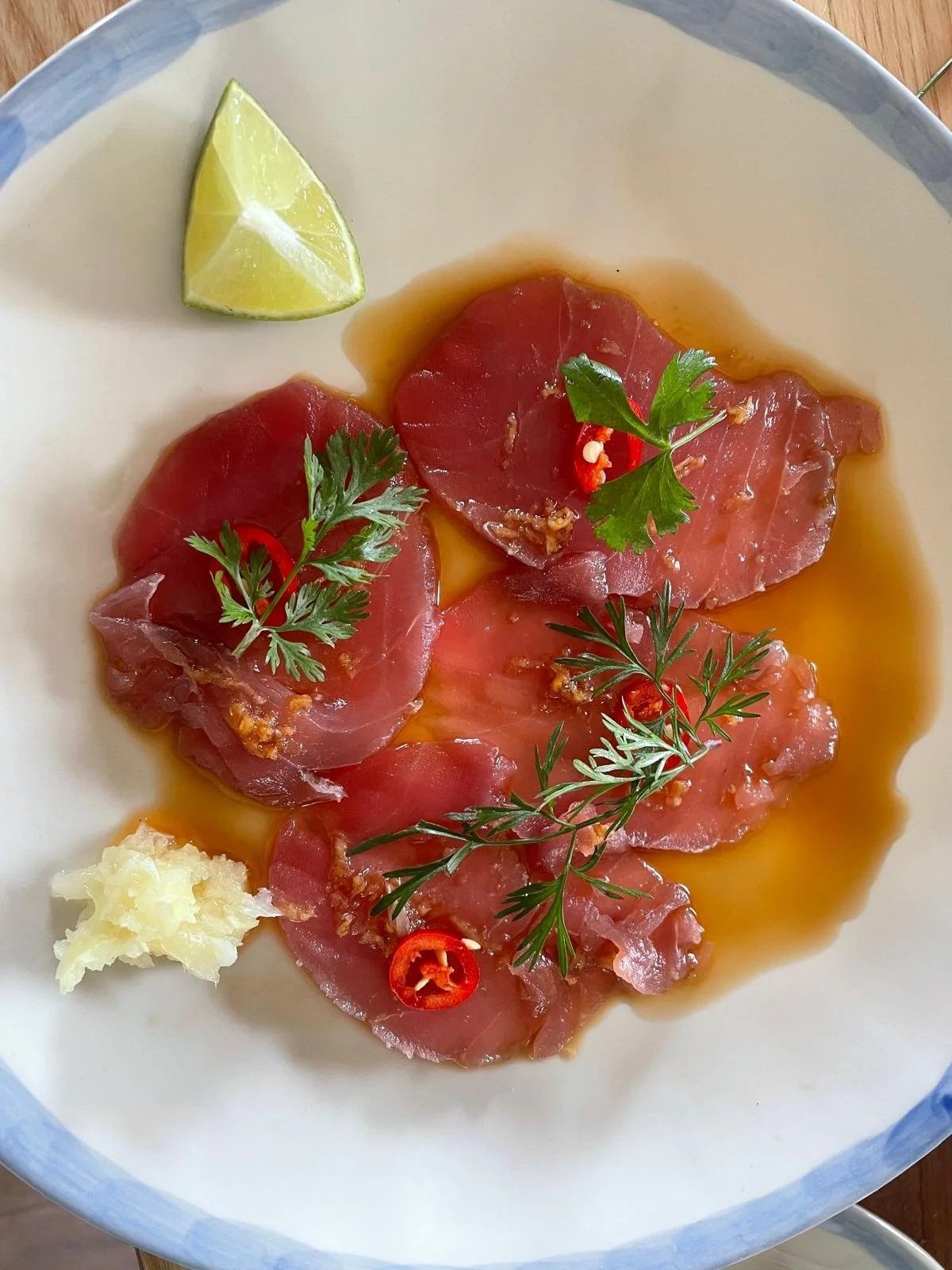 Ceviche with slices of raw fish, cilantro, red chili slices, a lime wedge, and garlic on a white plate.