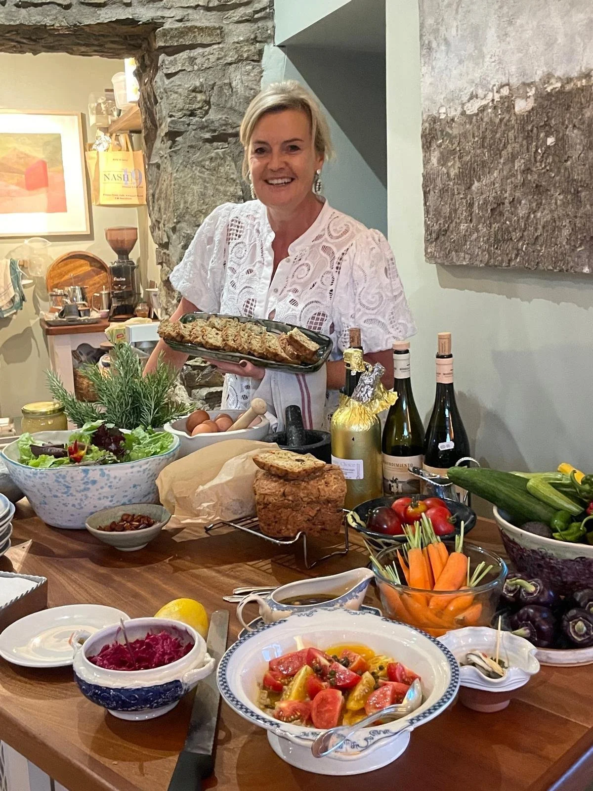 A woman smiling and holding a platter of food at a table filled with various dishes, vegetables, and bottles in a cozy, rustic kitchen.