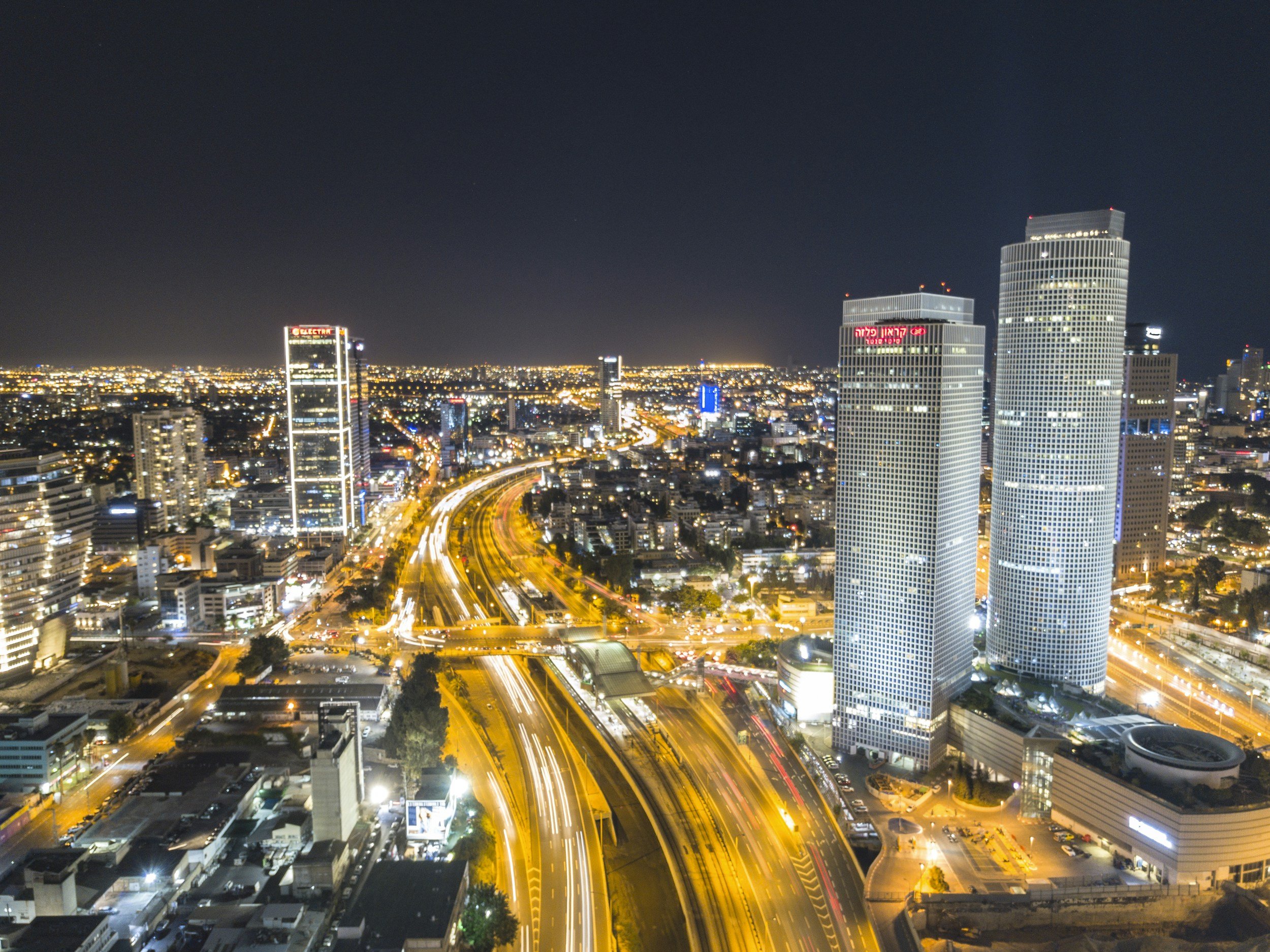 Nighttime cityscape of a densely built downtown area with tall skyscrapers, illuminated windows, and busy roads with streaks of car lights indicating movement.