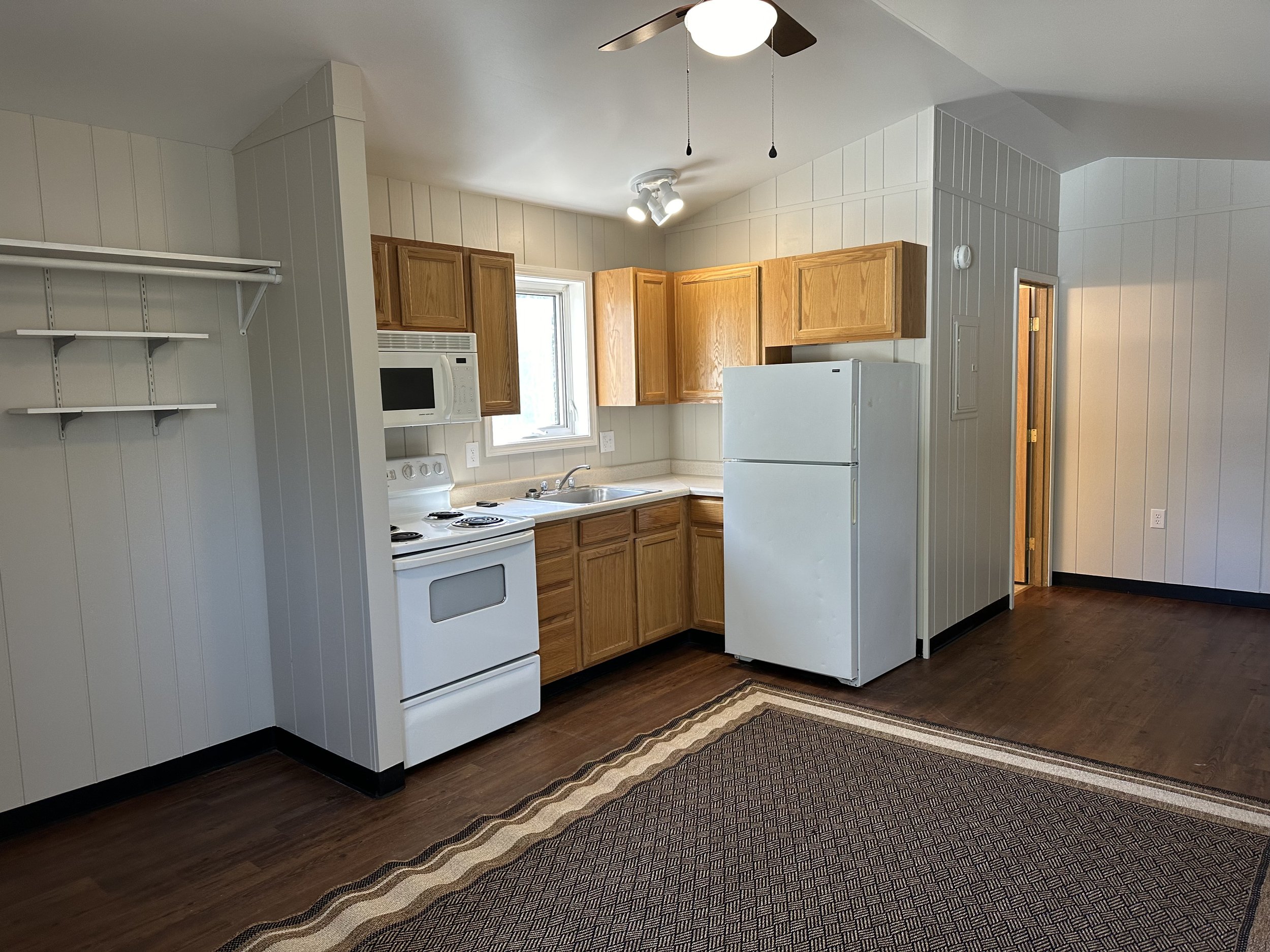 A small kitchen with white walls and wooden cabinets. It features a white stove, refrigerator, and microwave. There are shelves on a side wall, a ceiling fan, and a rug on hardwood flooring.
