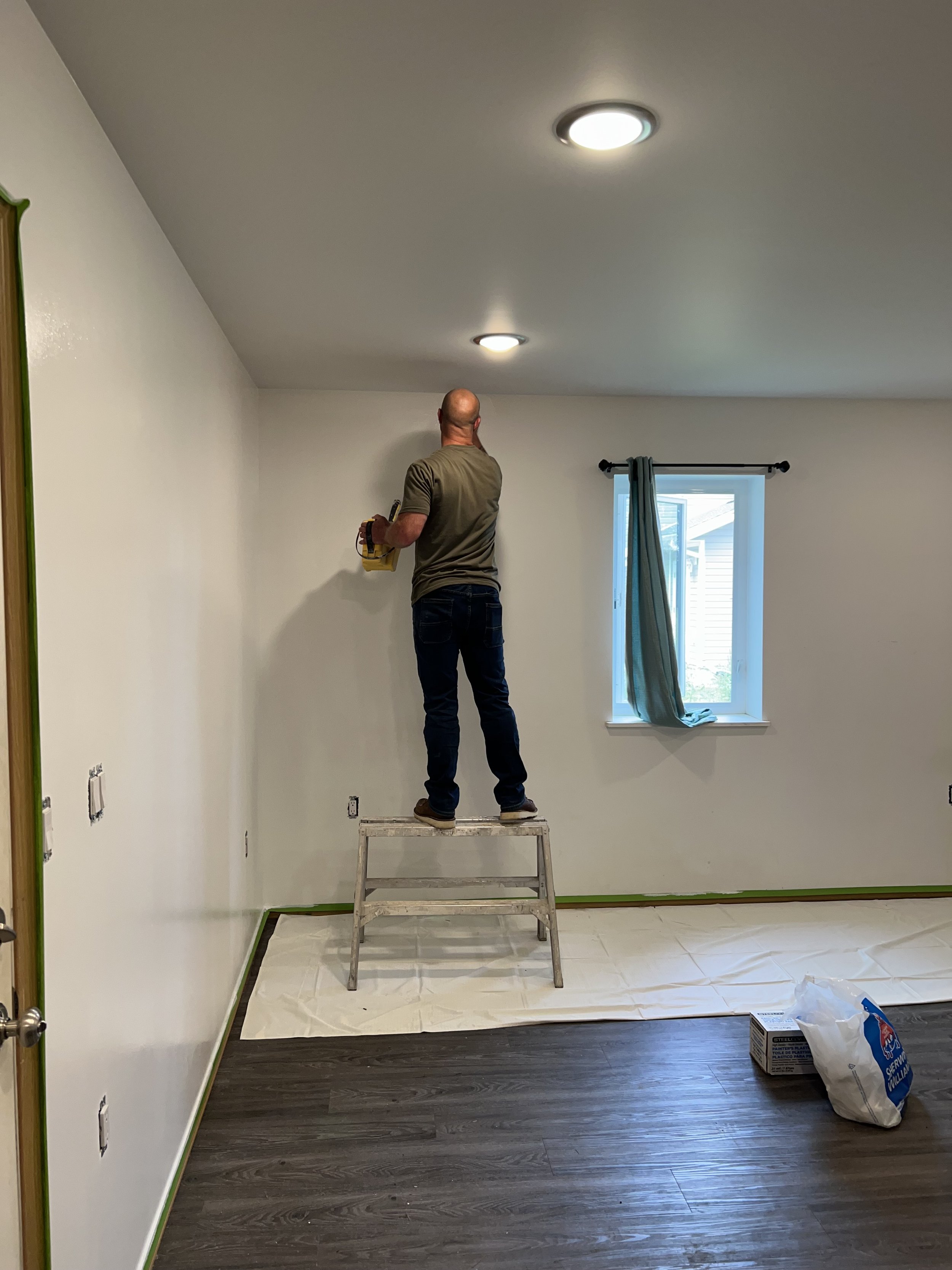 Person standing on ladder painting wall in a room with wooden floor and window with curtain.