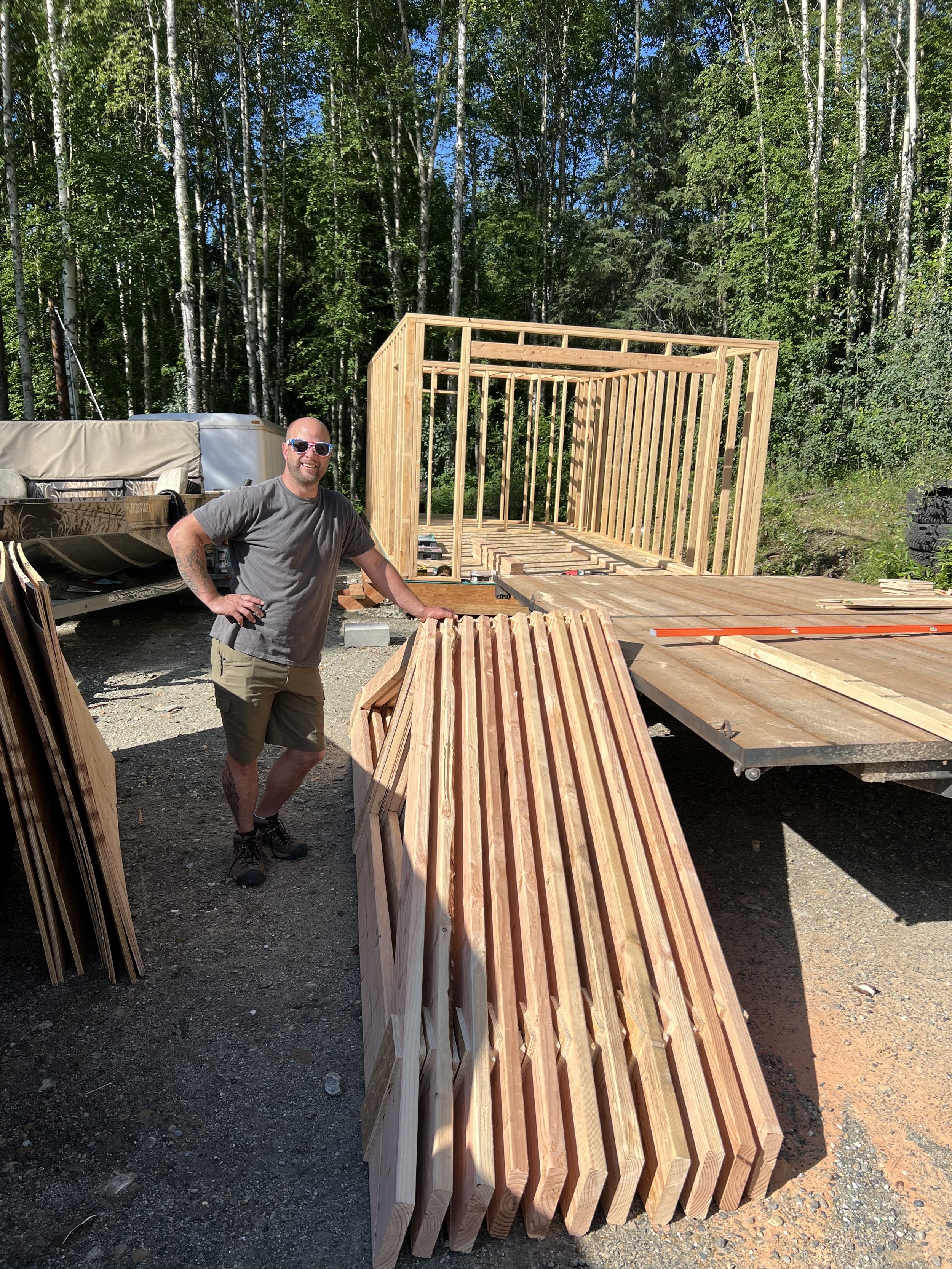 Person standing next to lumber and partially built wooden structure in outdoor setting with trees.