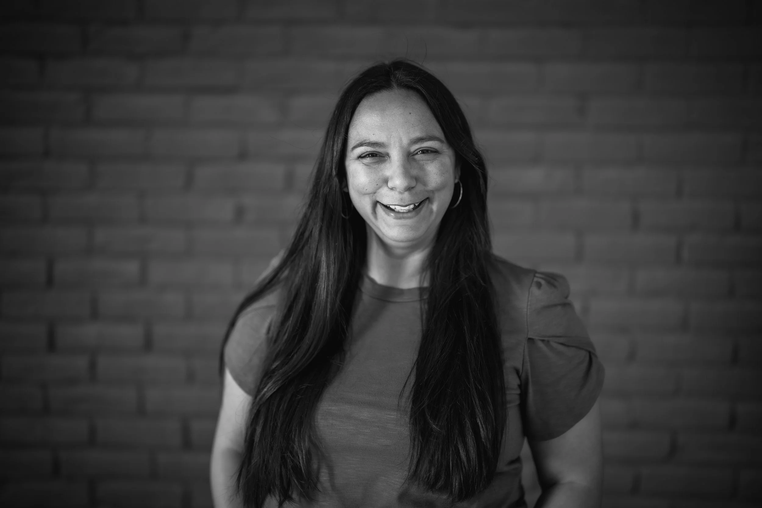 A person in a black shirt with a praying hands logo stands indoors, smiling slightly, with a plant and a curtain in the background.