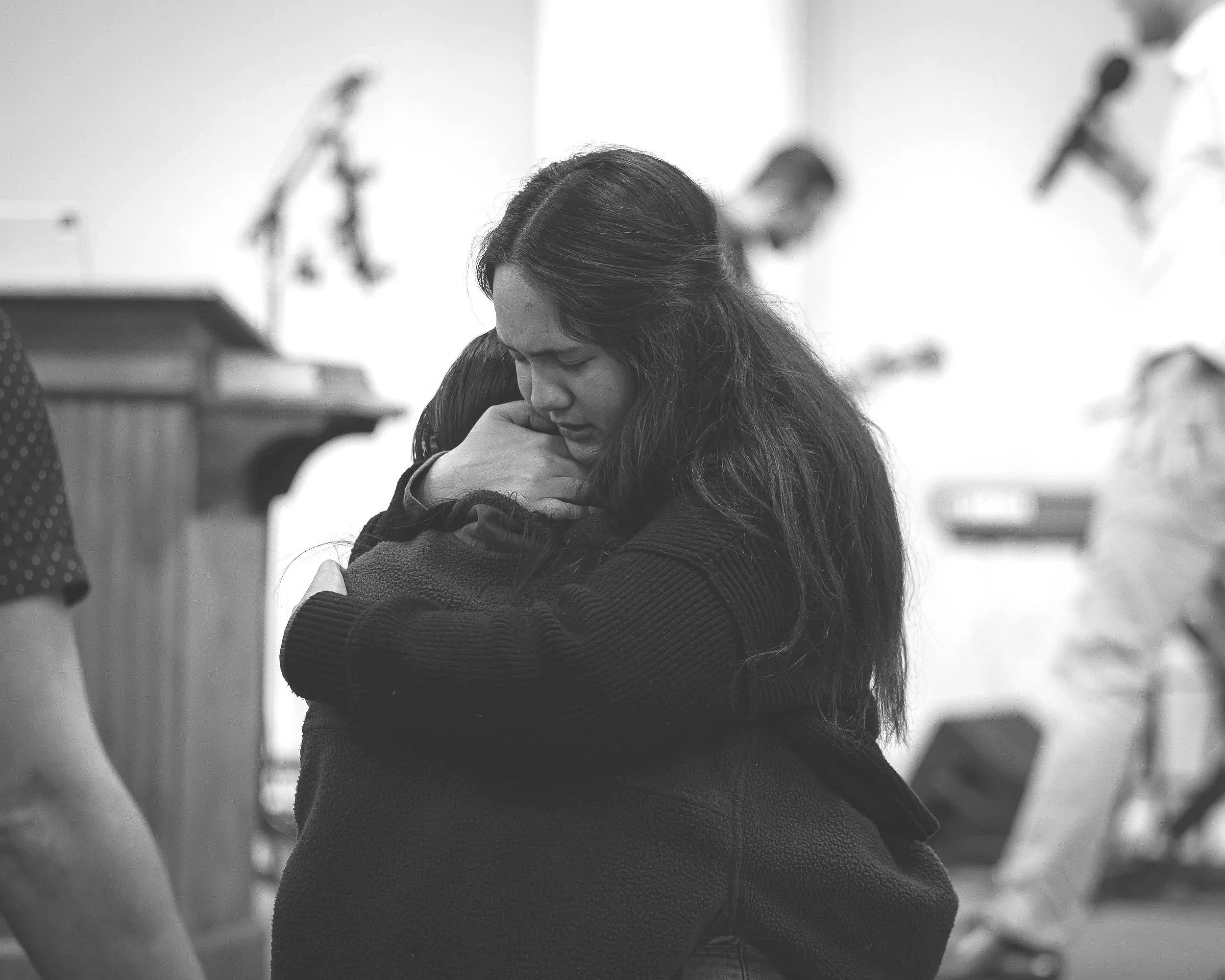 A woman hugging a young girl in black clothing in a room with musical instruments in the background.