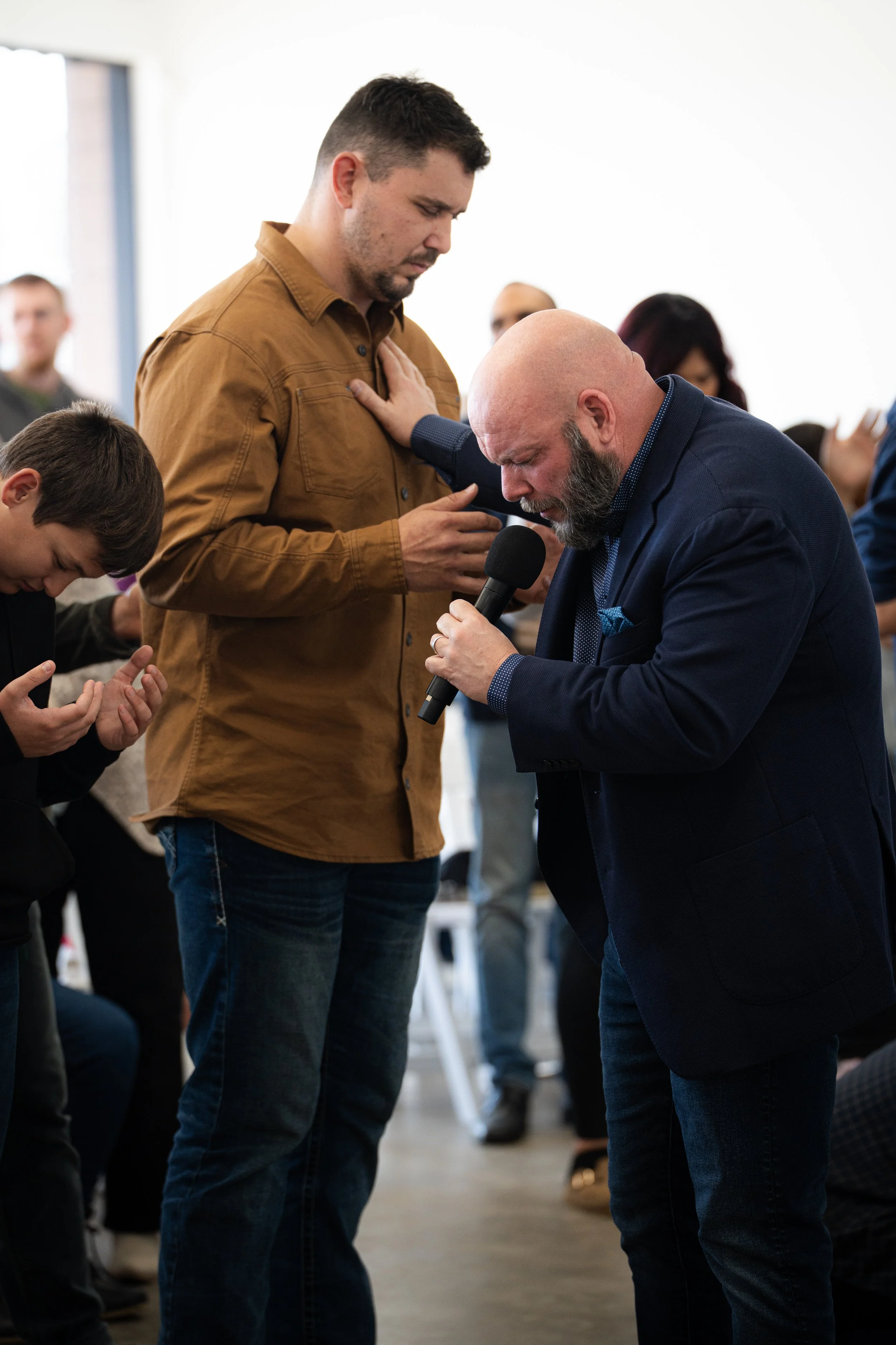 A man praying with a microphone, placing a hand on another man's chest, while people around have heads bowed in a prayerful setting.