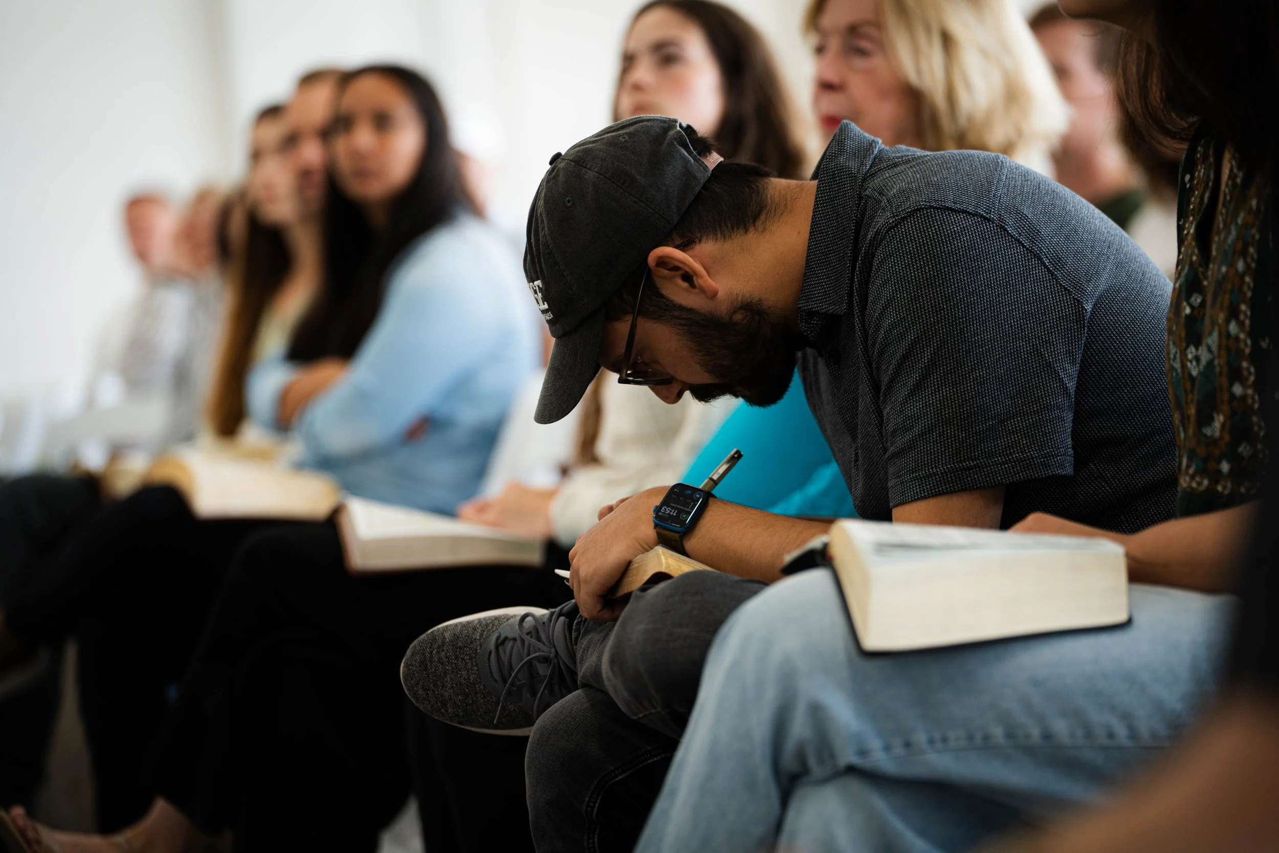 A man wearing a black cap and wristwatch takes notes during a lecture while sitting among a diverse group of students with books.