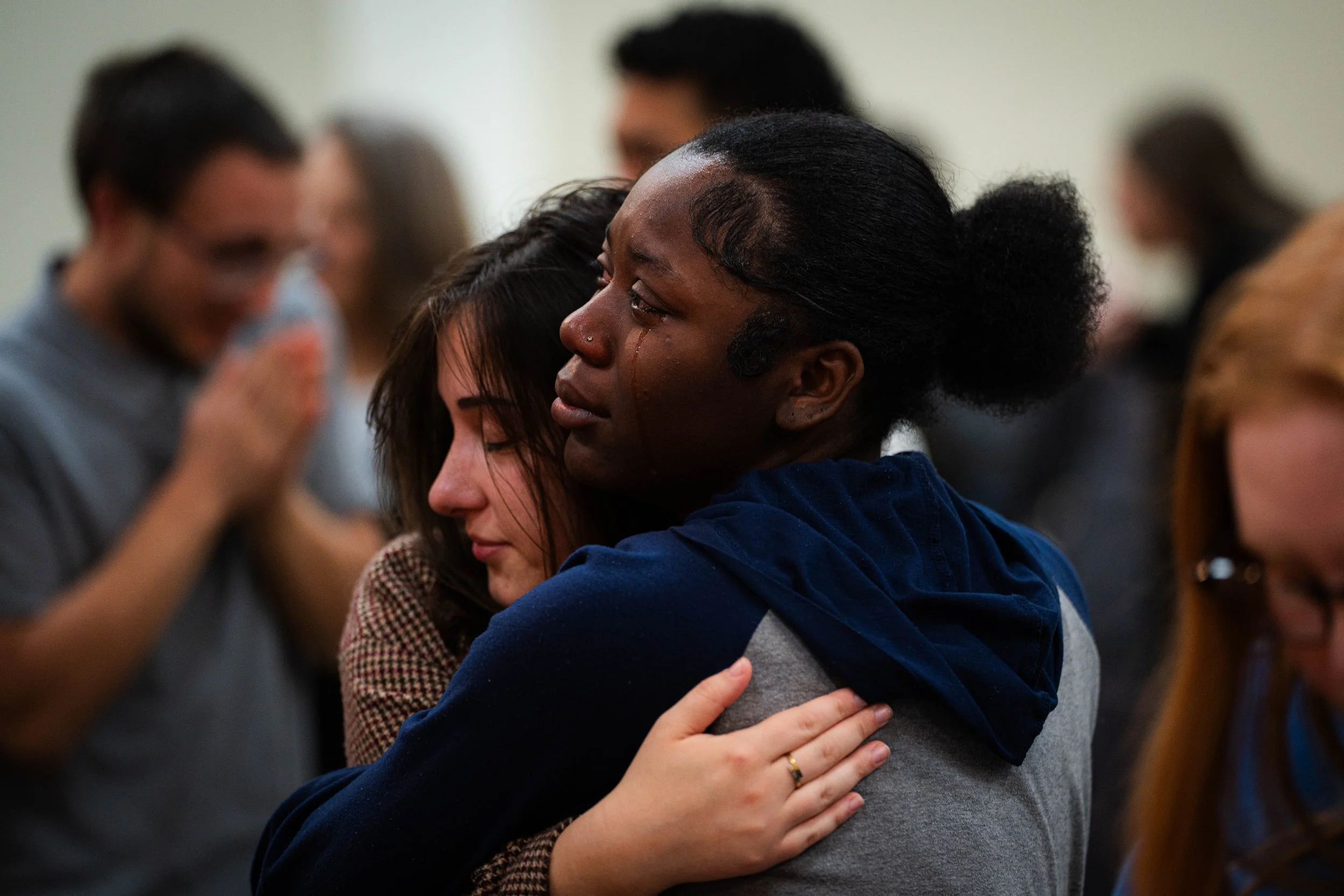 Two women hugging, with one crying, surrounded by other people in a somber gathering.
