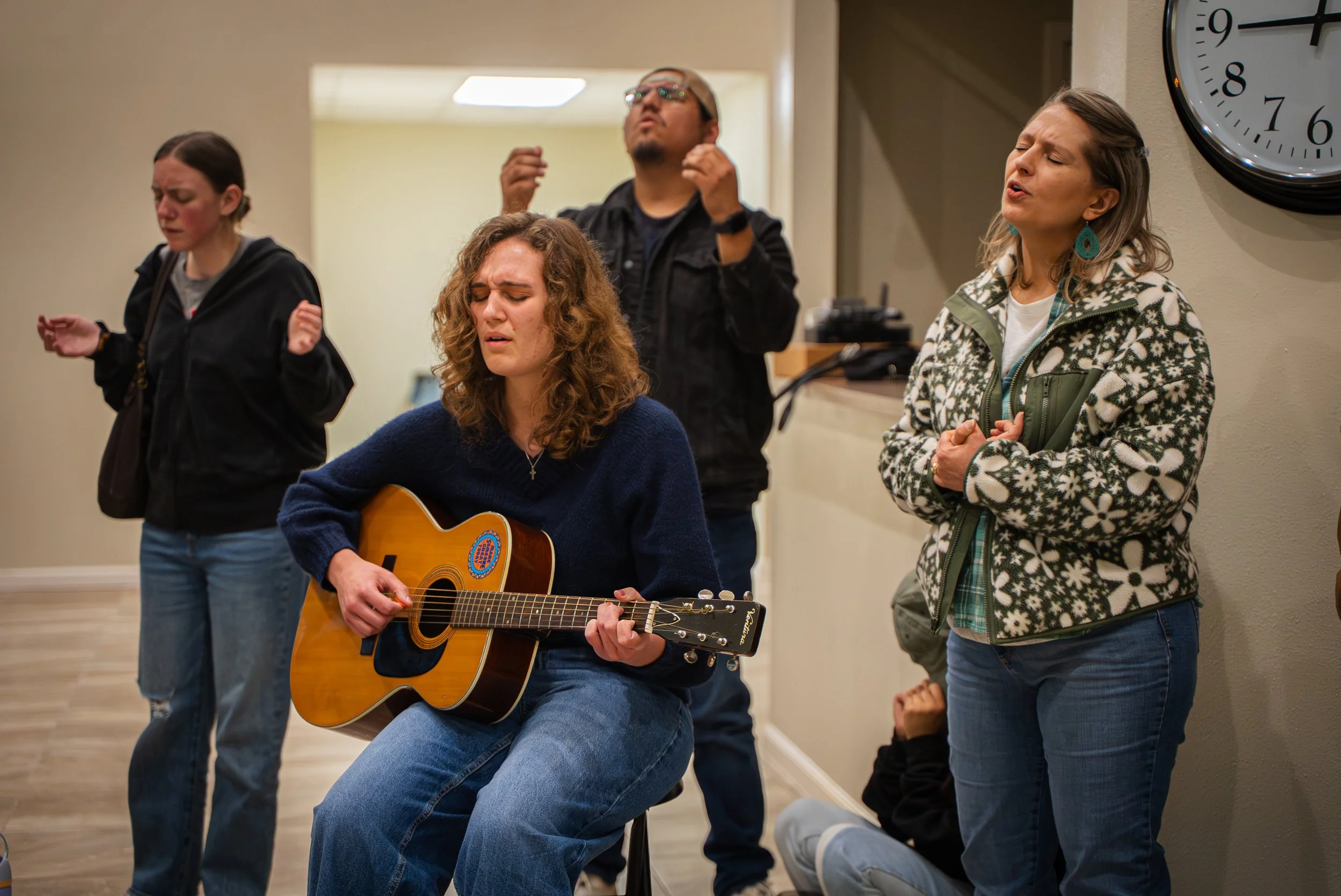 Group of people singing and playing guitar in a room, with a clock on the wall.