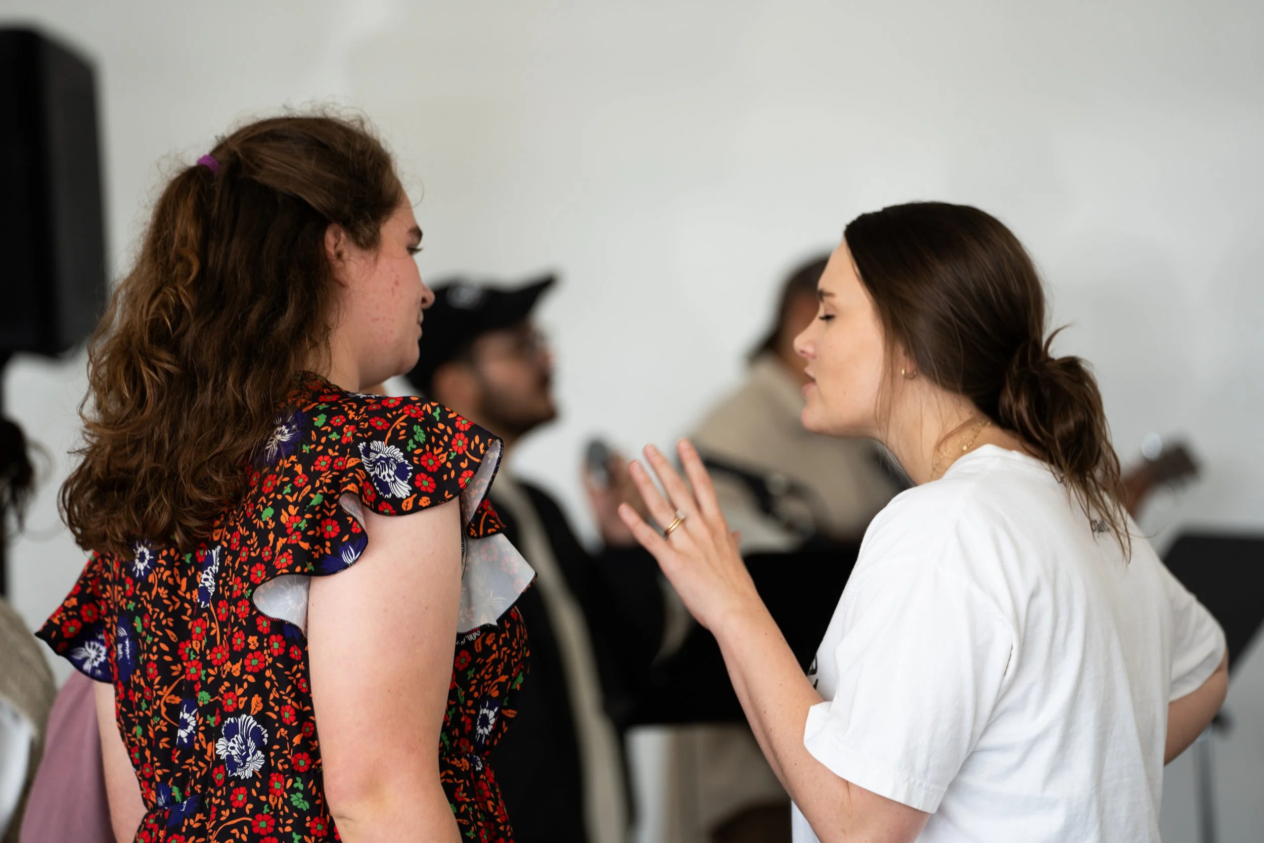 Two women having an intense conversation indoors, with a woman in white gesturing with her hand towards the woman with curly hair dressed in a floral dress.