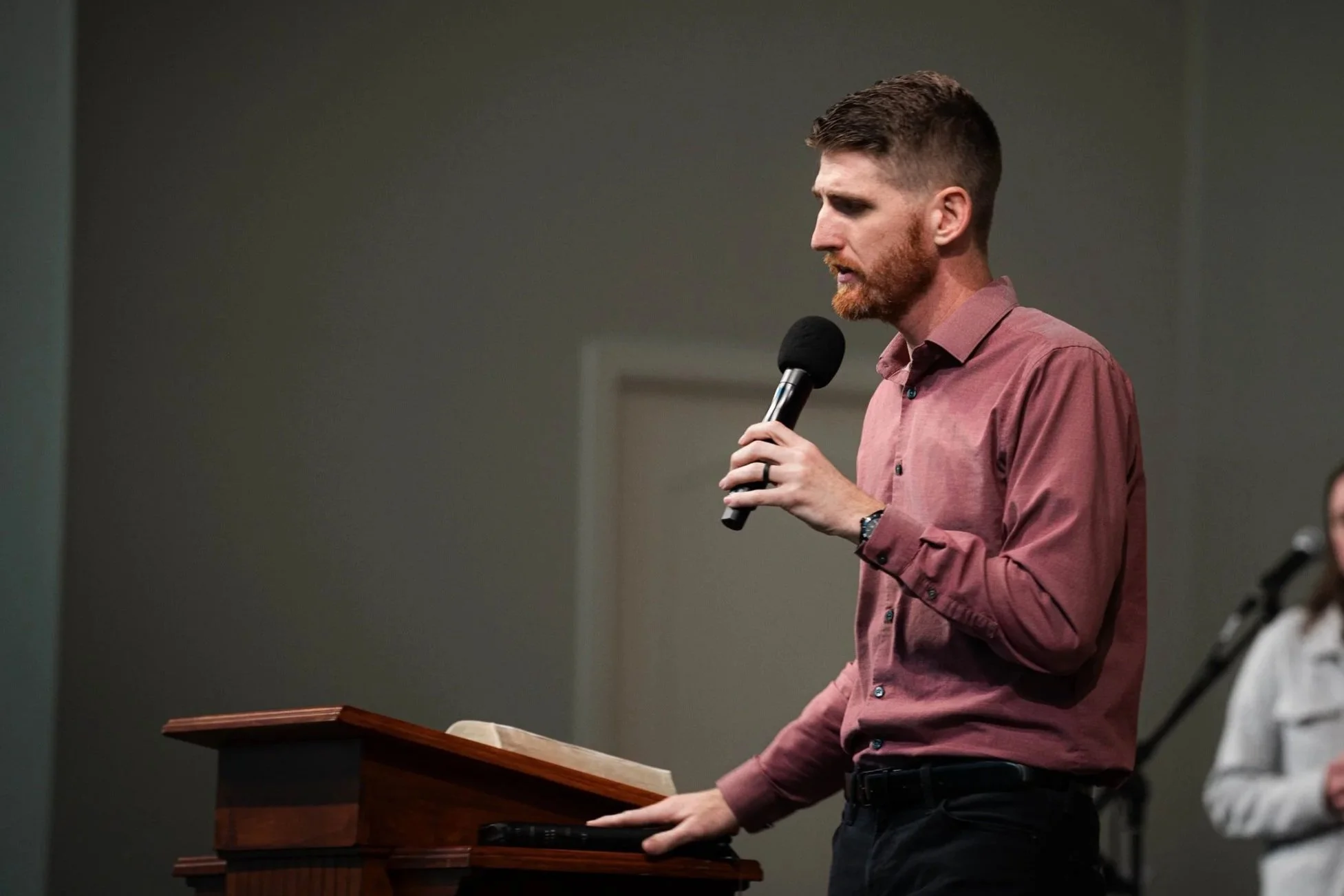 A man with short brown hair, beard, wearing a pink long sleeve shirt, holding a microphone and speaking at a podium.