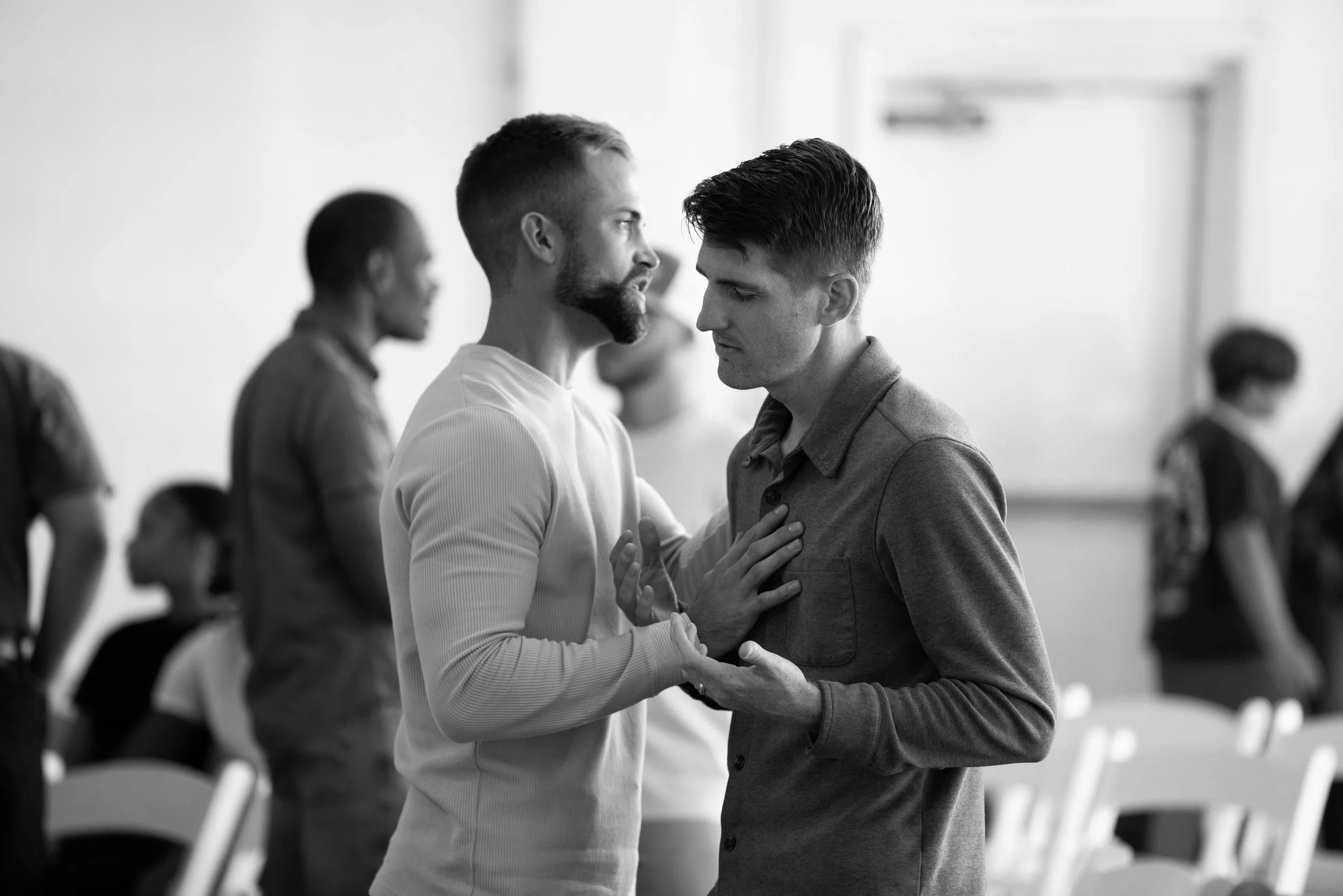 Two men in a church or religious setting, with one holding the other’s chest in a prayer position, surrounded by other people, some praying and some standing.