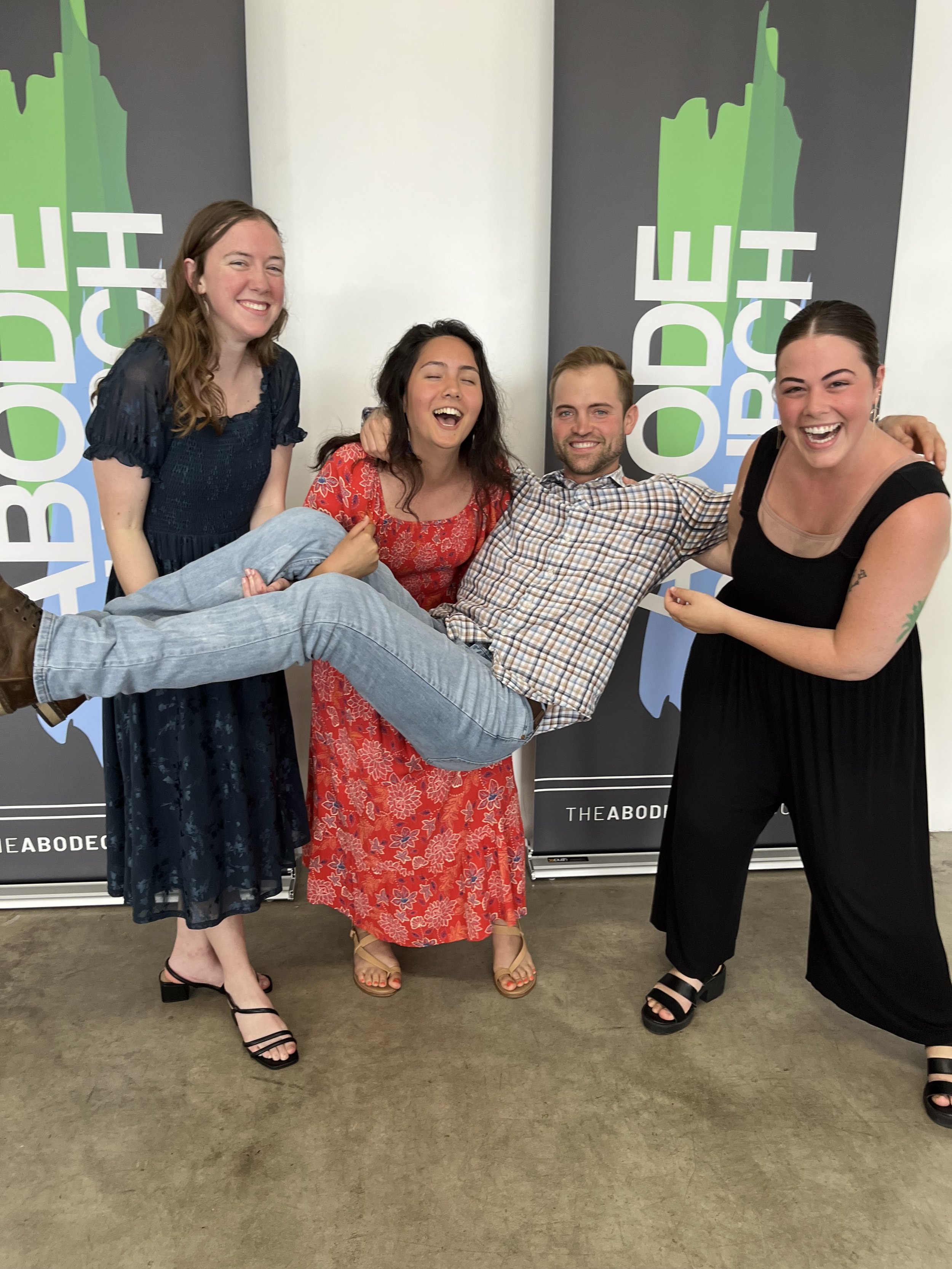 Three women holding a man horizontally in front of a backdrop with text "THE ABODE CHURCH". They are all smiling and appear to be having fun.