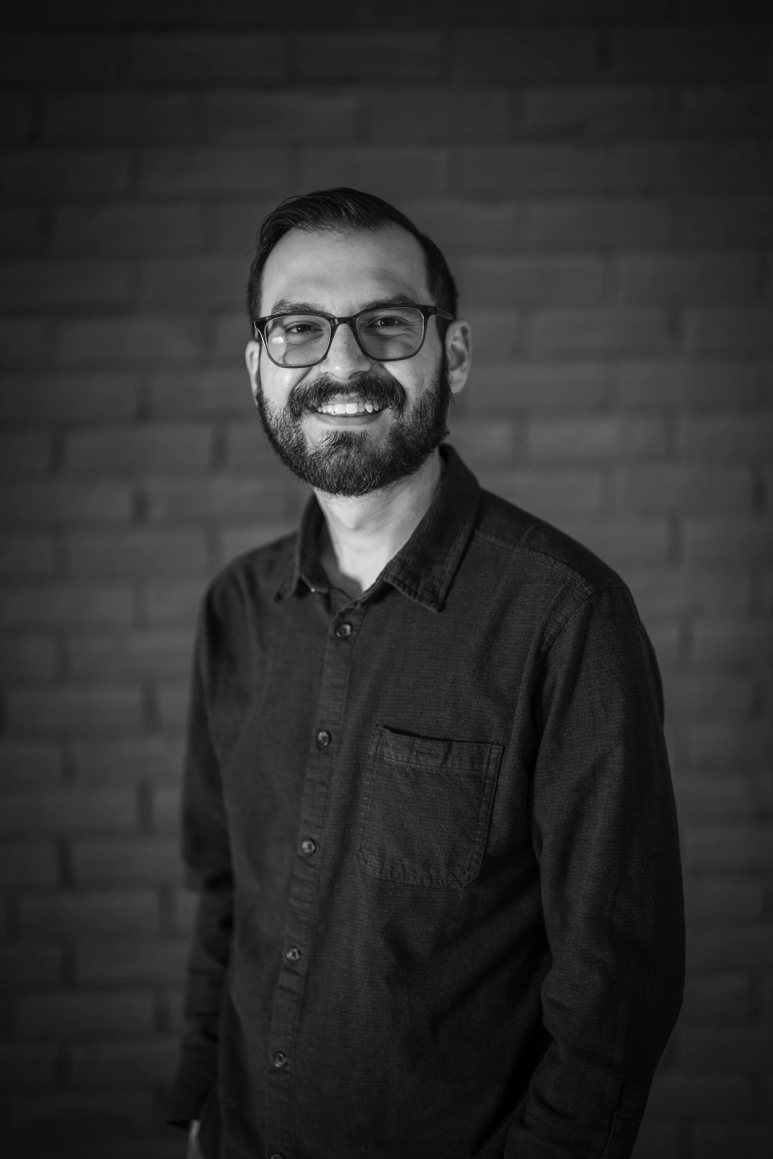 Black and white photo of a smiling man with glasses and a beard, wearing a dark button-up shirt, standing in front of a brick wall.
