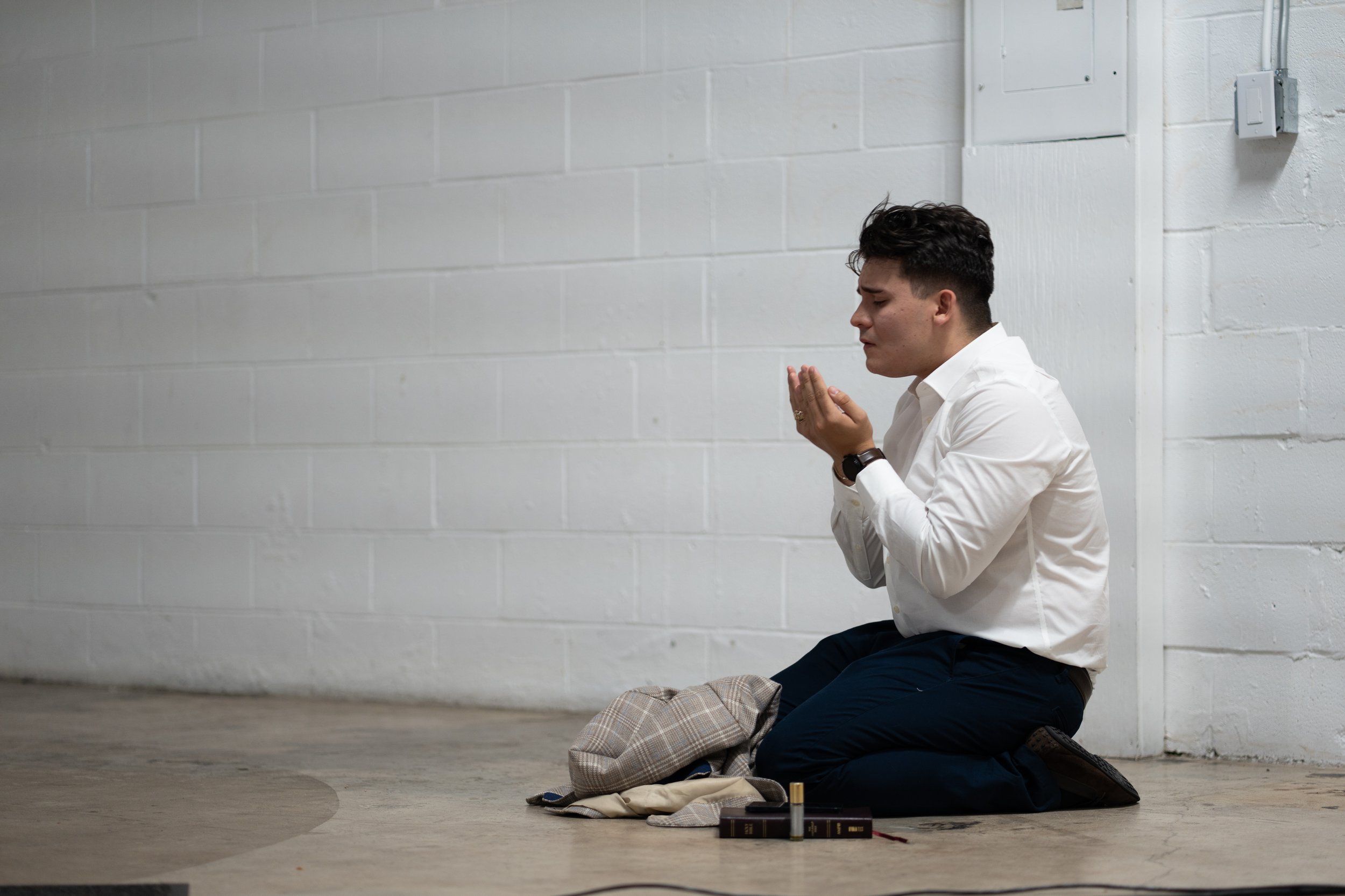 Person in white shirt and dark pants kneeling on the floor in prayer, hands raised, against a light gray brick wall.