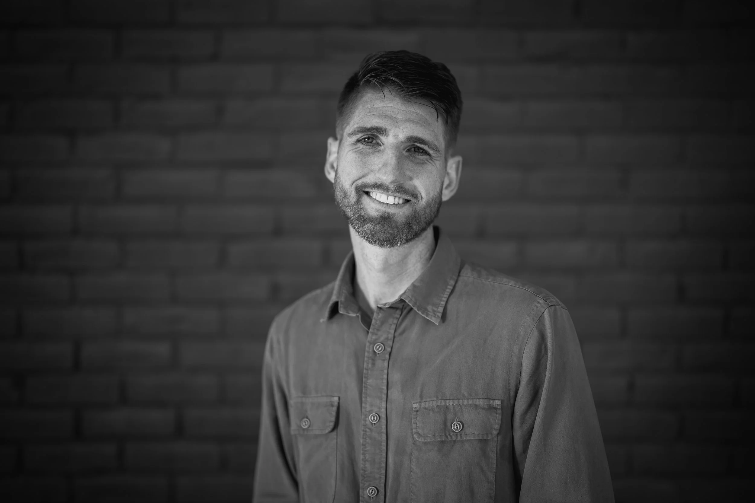 A young man with red hair and a beard, wearing a dark blazer and black shirt, sitting on a stool in a dimly lit room with modern decor.