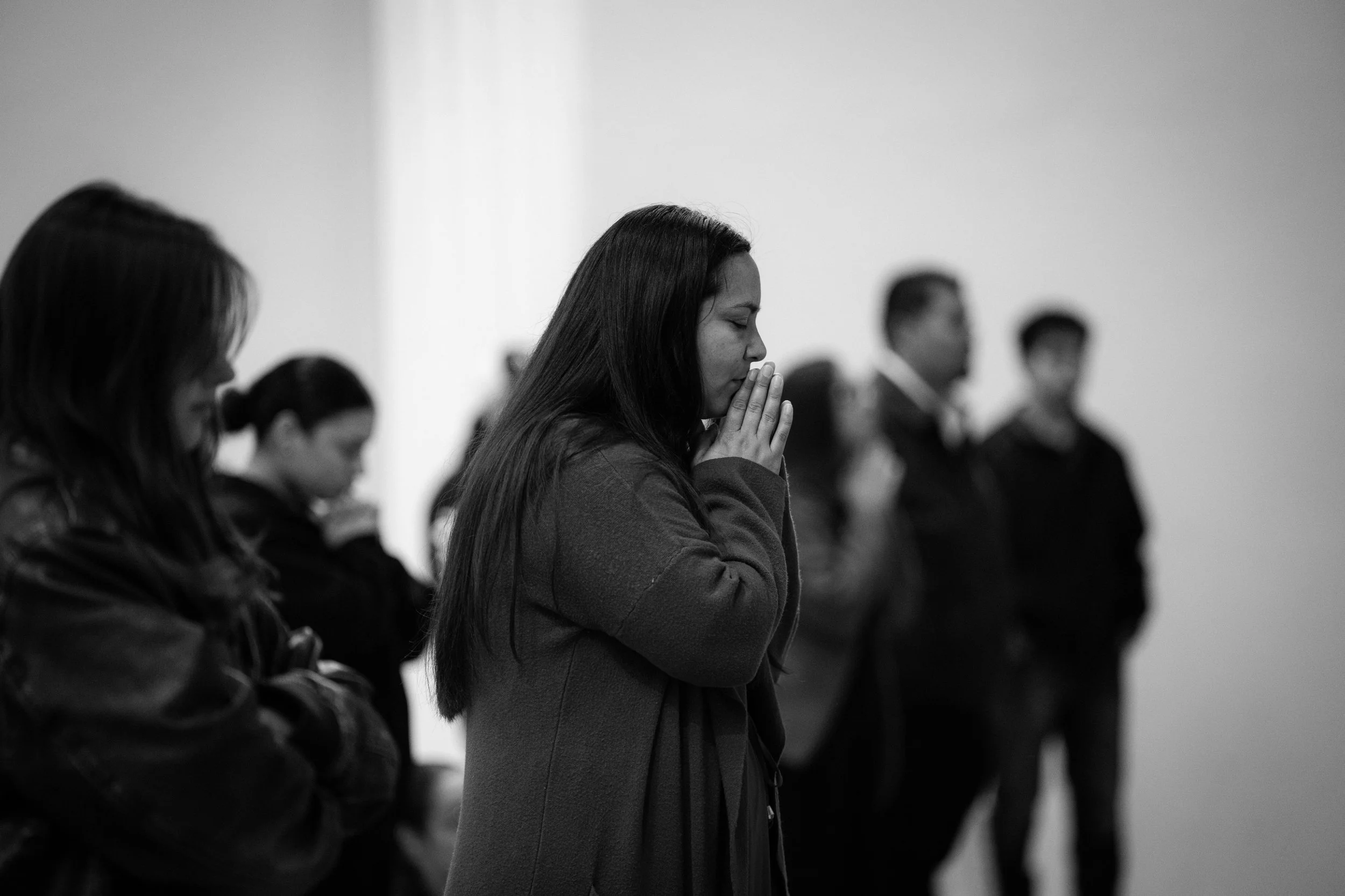 A group of people, mostly women, standing with their eyes closed and hands clasped in prayer in a room with plain walls.