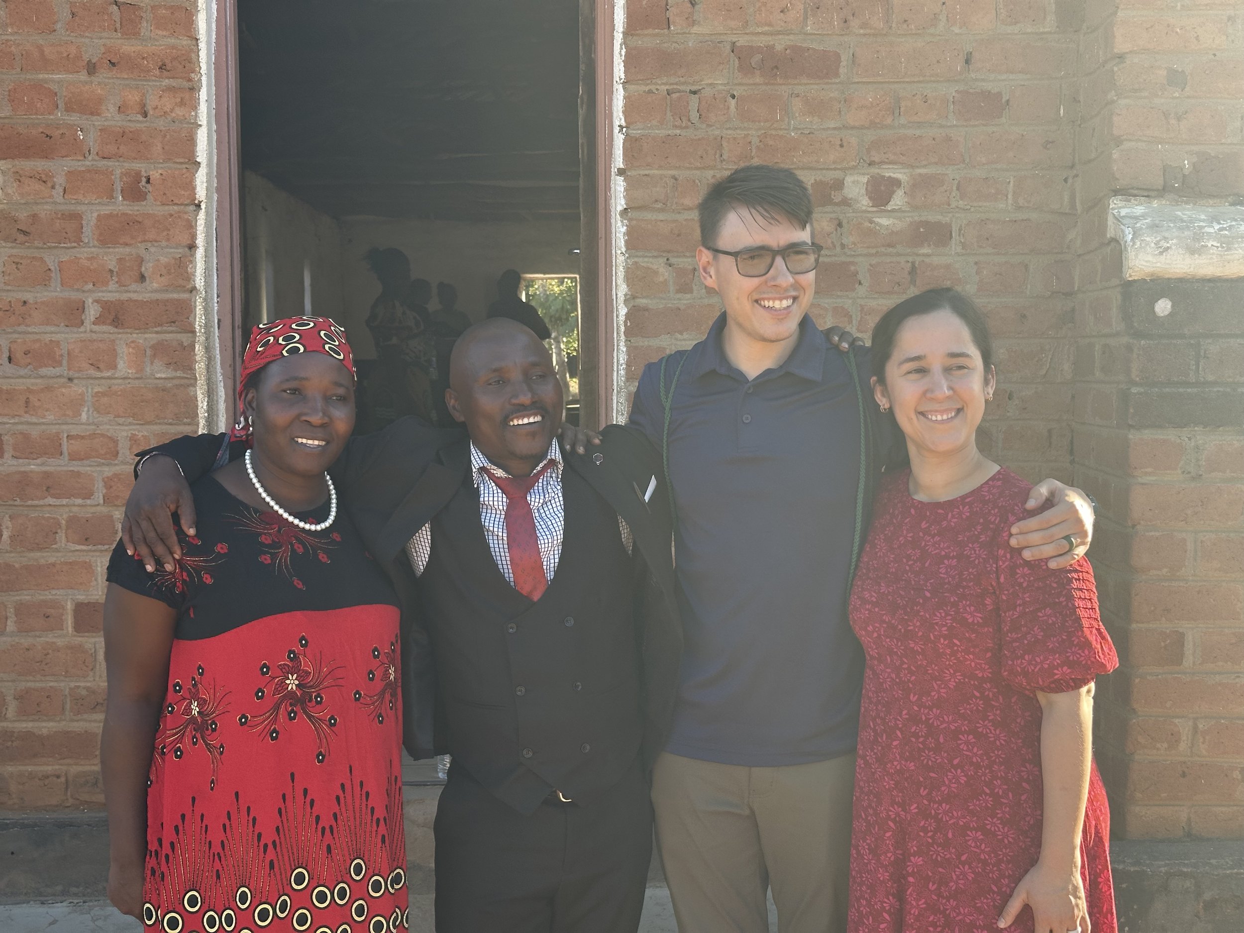 Four people posing for a photo in front of a brick building. The group includes three individuals wearing dressed up attire and one person in a casual polo shirt. One person is wearing a colorful headscarf and dress, while another is in a suit and ti