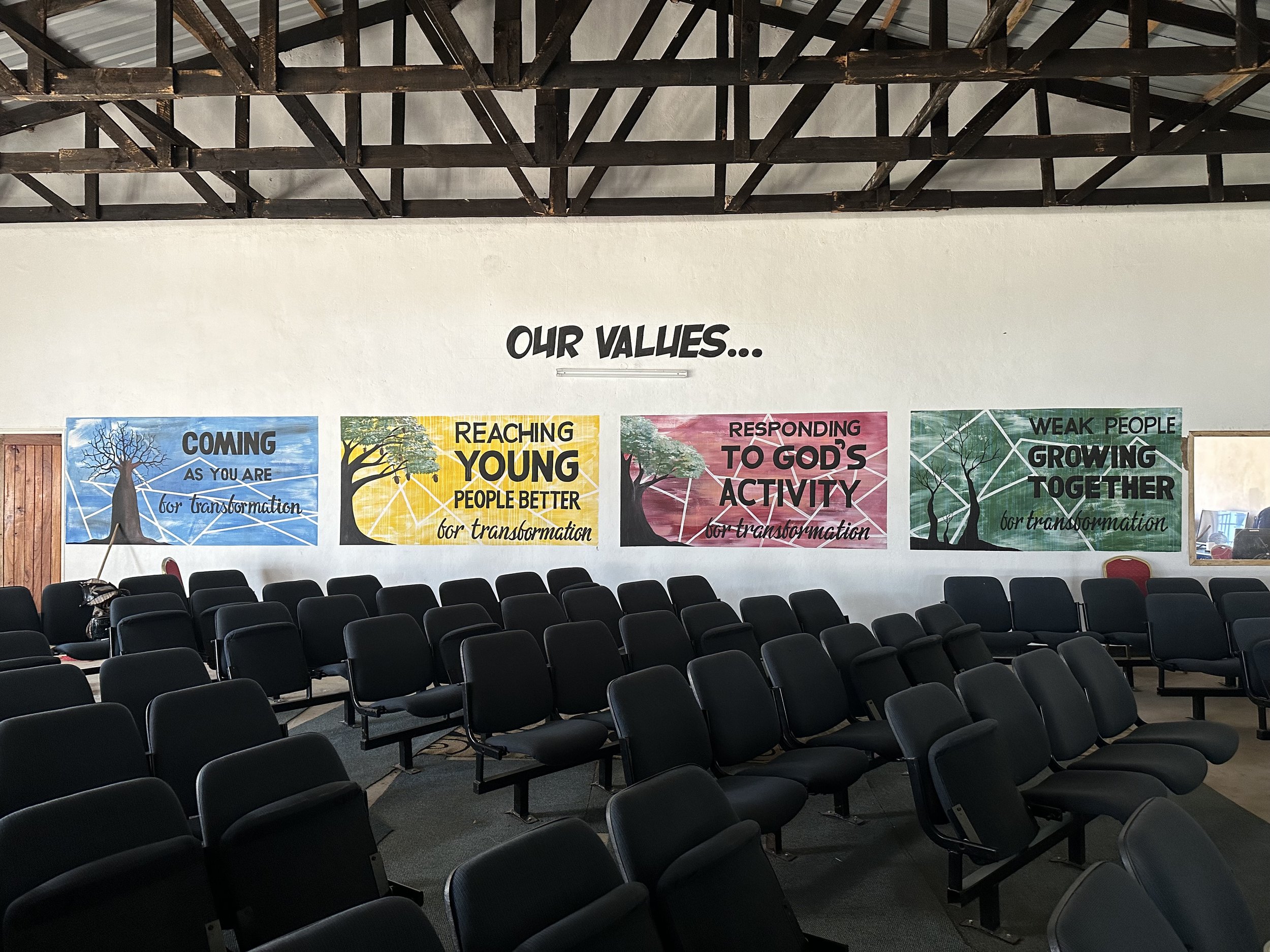 Interior of a room with rows of empty chairs and four colorful banners on the wall displaying values related to transformation, spiritual growth, and community involvement.