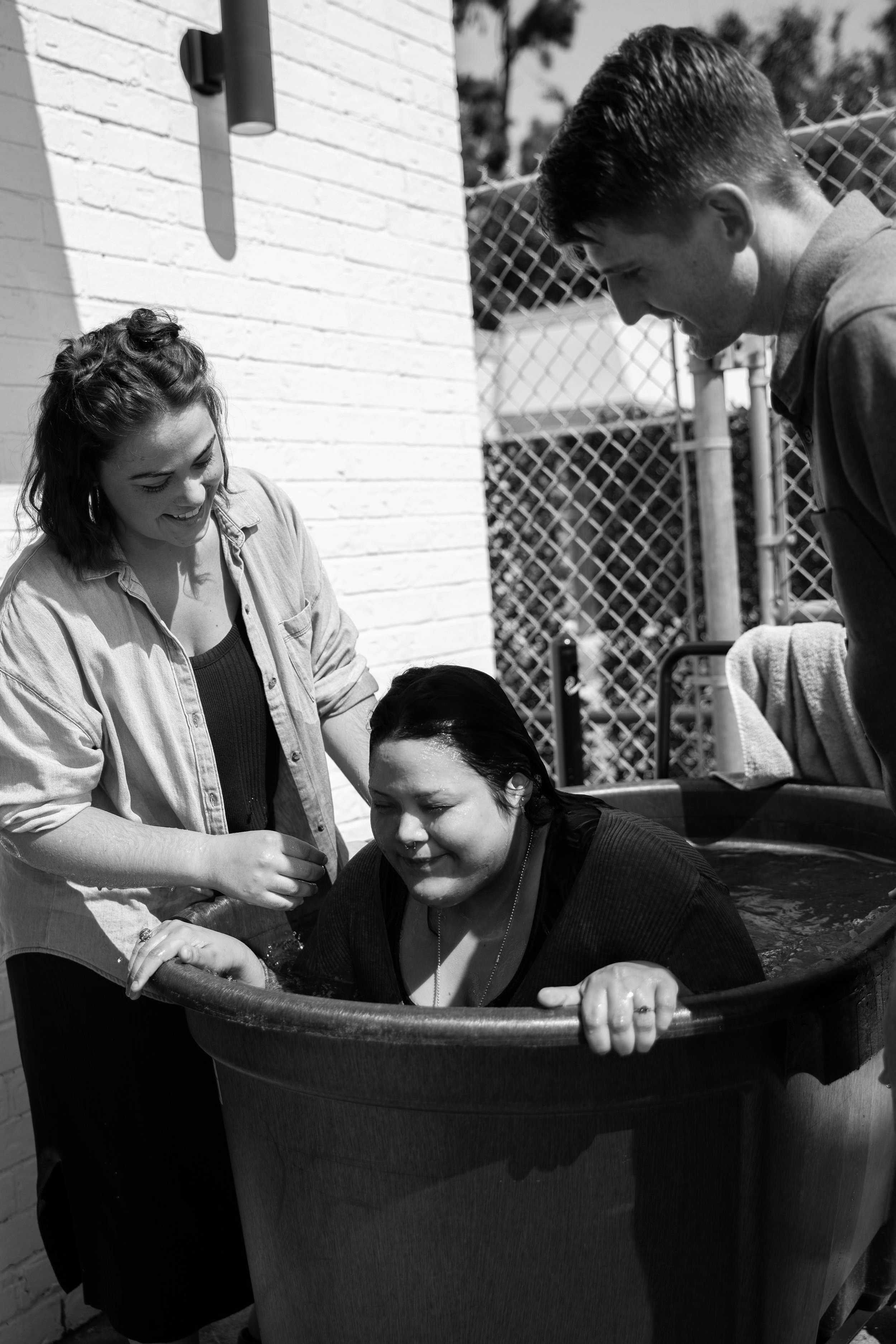 A woman in a baptism tub supported by two people outdoors beside a brick wall and chain-link fence.