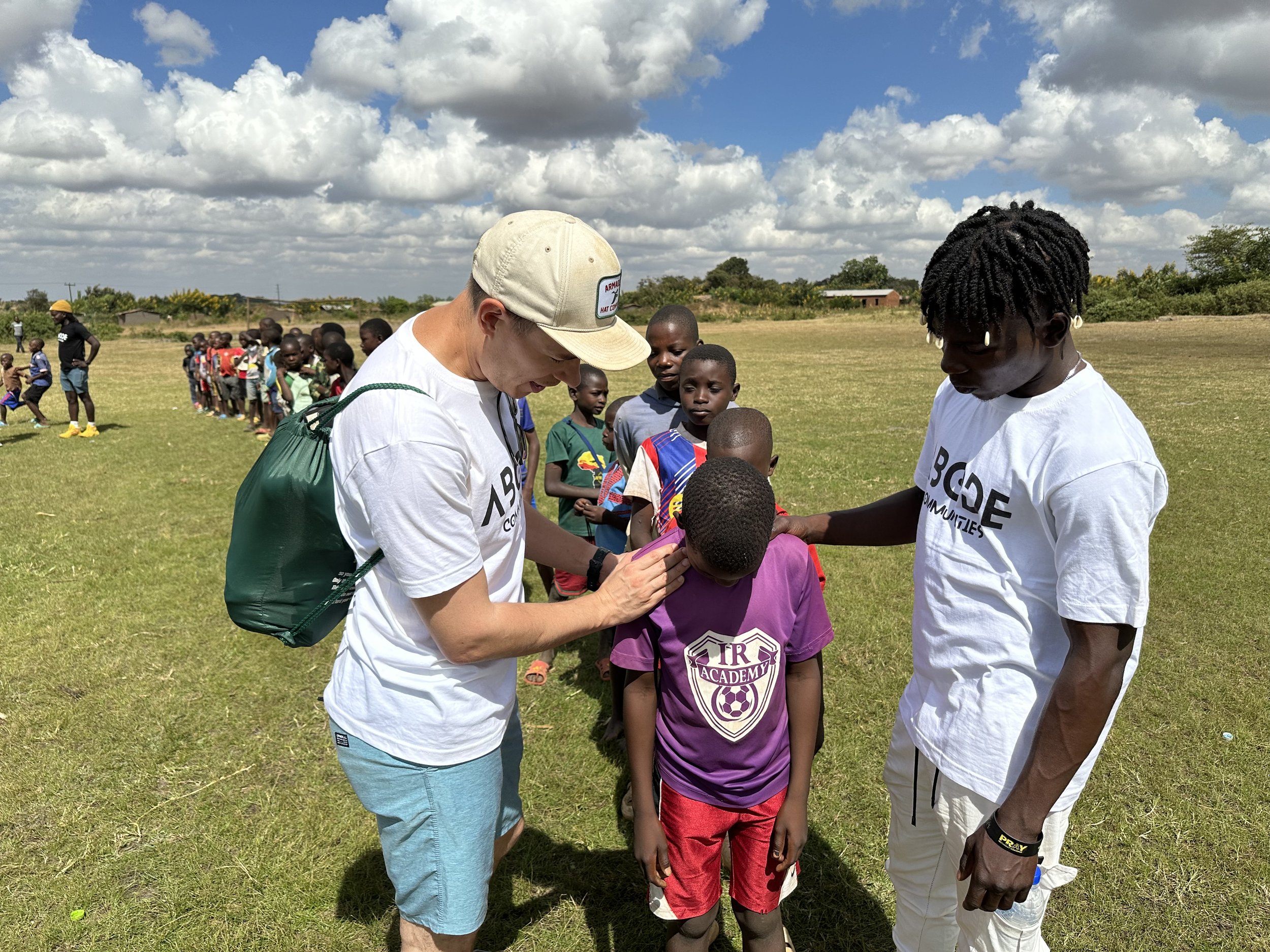 Two adults interacting with a child in a sports field; one adult writing or adjusting something on the child's shirt, and another adult standing nearby. A group of children is lined up in the background on the grassy field with a partly cloudy sky ov