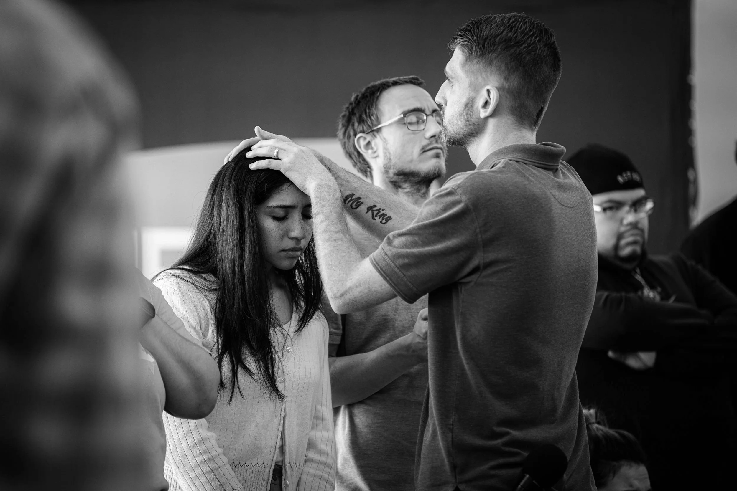 People engaged in a prayer session, with a man laying hands on a woman's head, eyes closed, in a solemn setting.